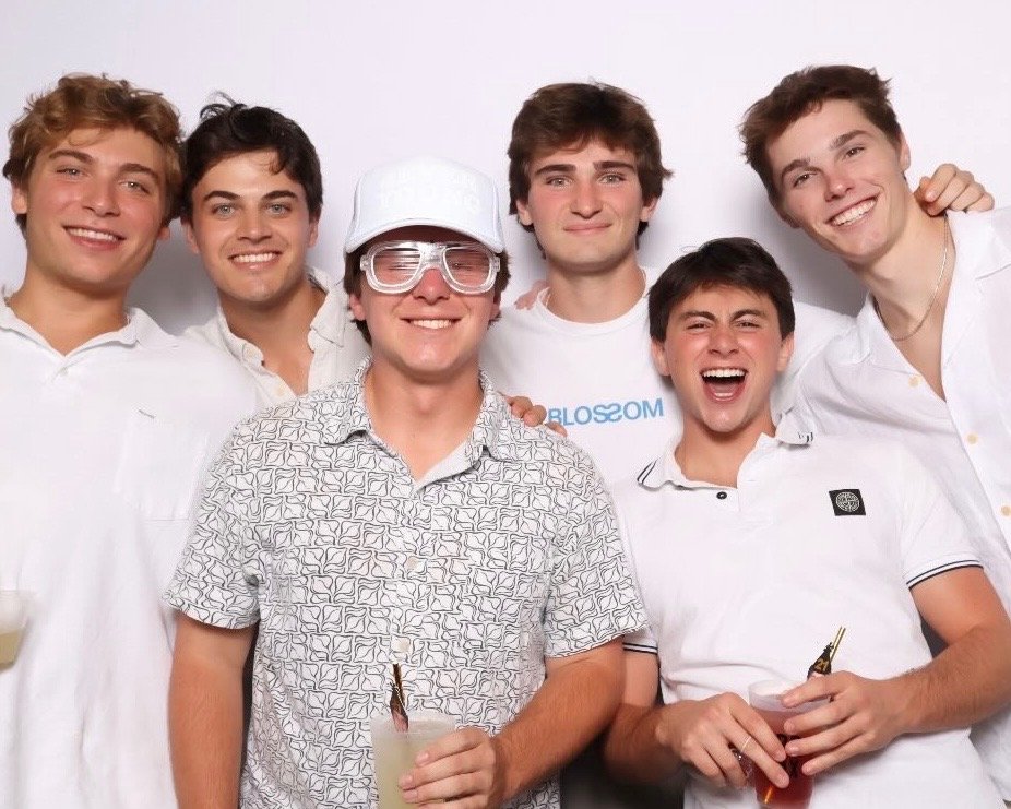 Group of six young men smiling and posing together at a party, some holding drinks, indoors against a plain white background.