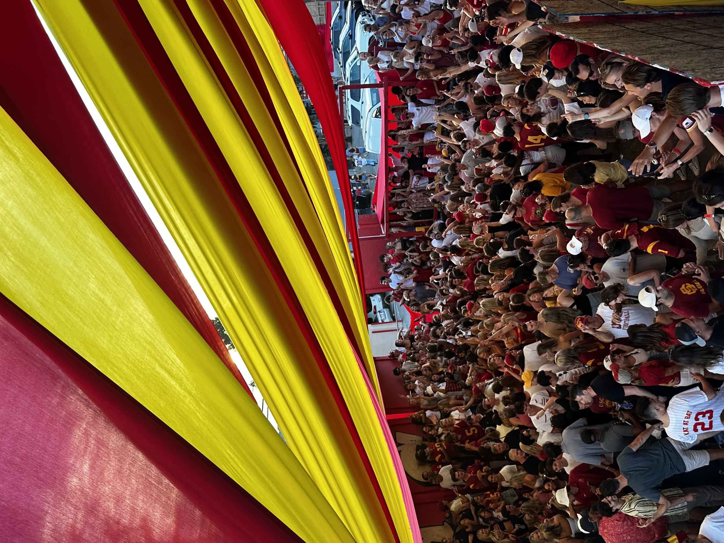 Crowd of people, many in red and yellow clothing, gathered at a sports event under a covered area, with a row of parked cars visible in the background.