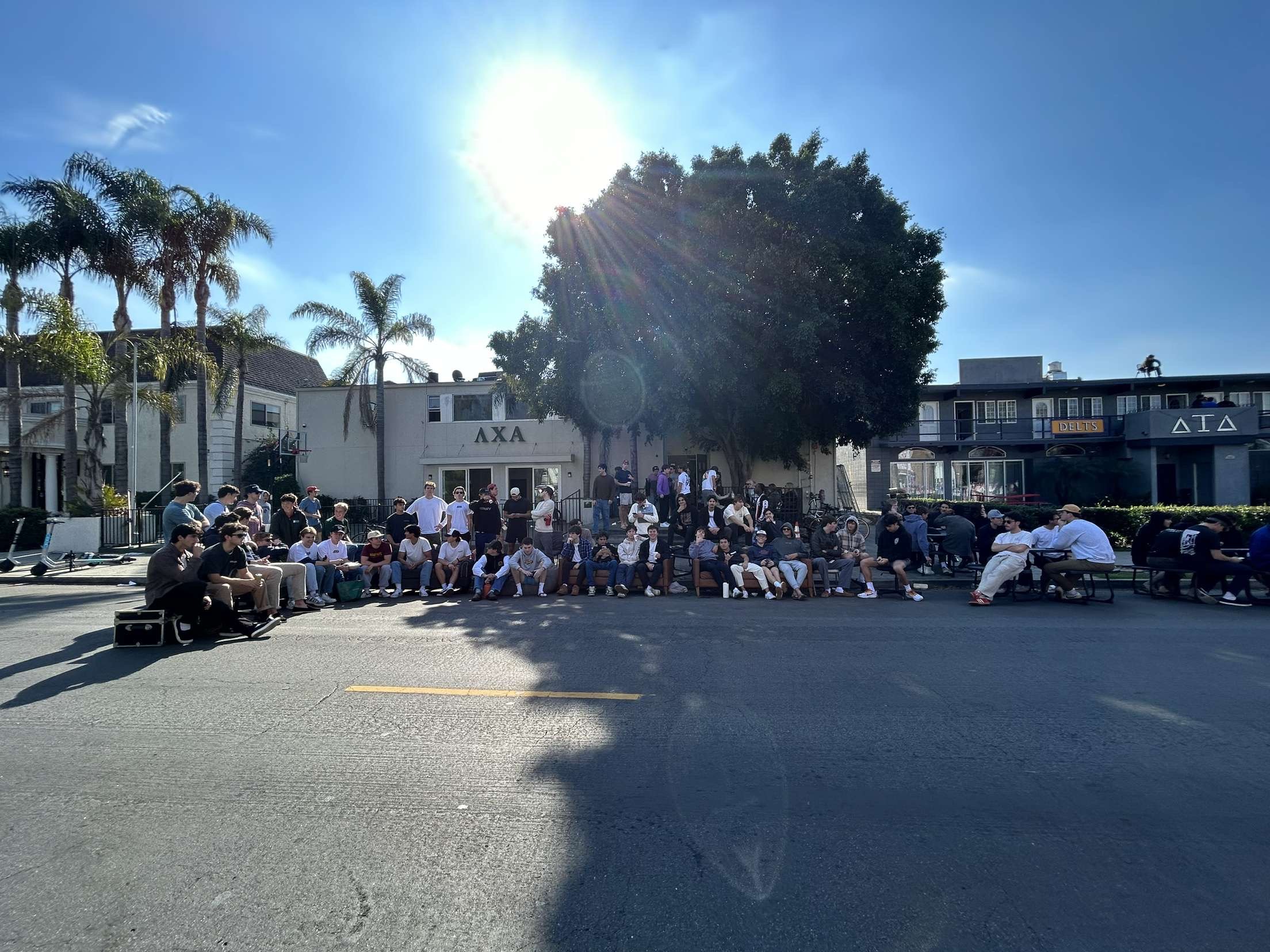 A large group of young people gathered outdoors on a sunny day, sitting on benches and standing, with palm trees and buildings in the background.