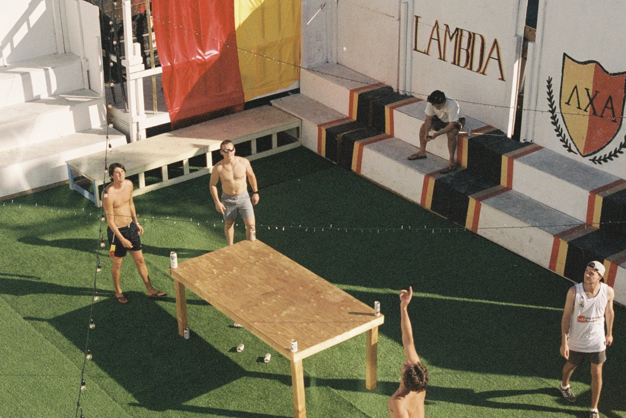 People playing a game of beer pong outdoors on a grassy area near a stage with steps and a shield logo with Greek letters.