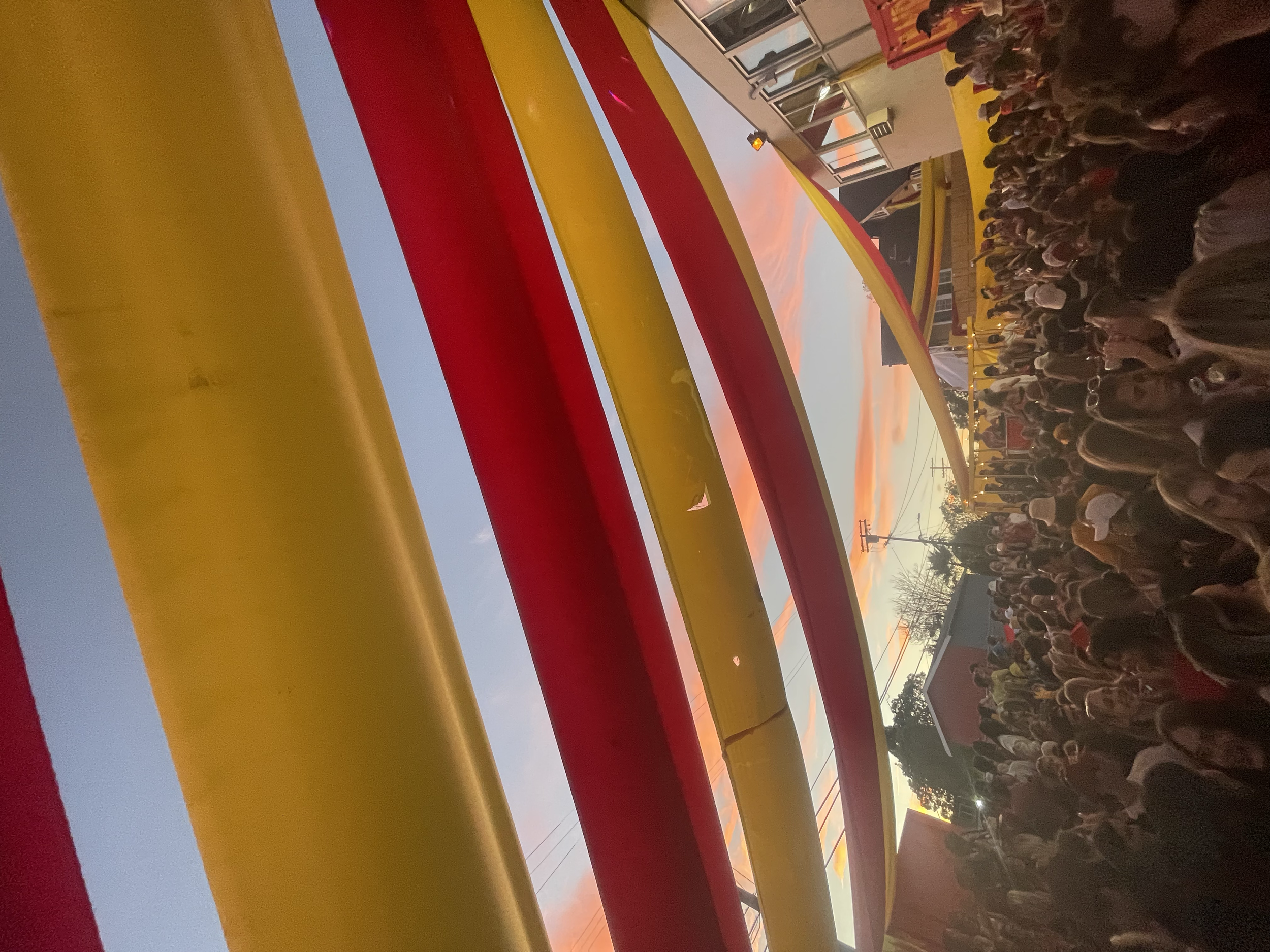 Crowd of people gathered outdoors during sunset, viewed from behind colorful yellow and red railings.