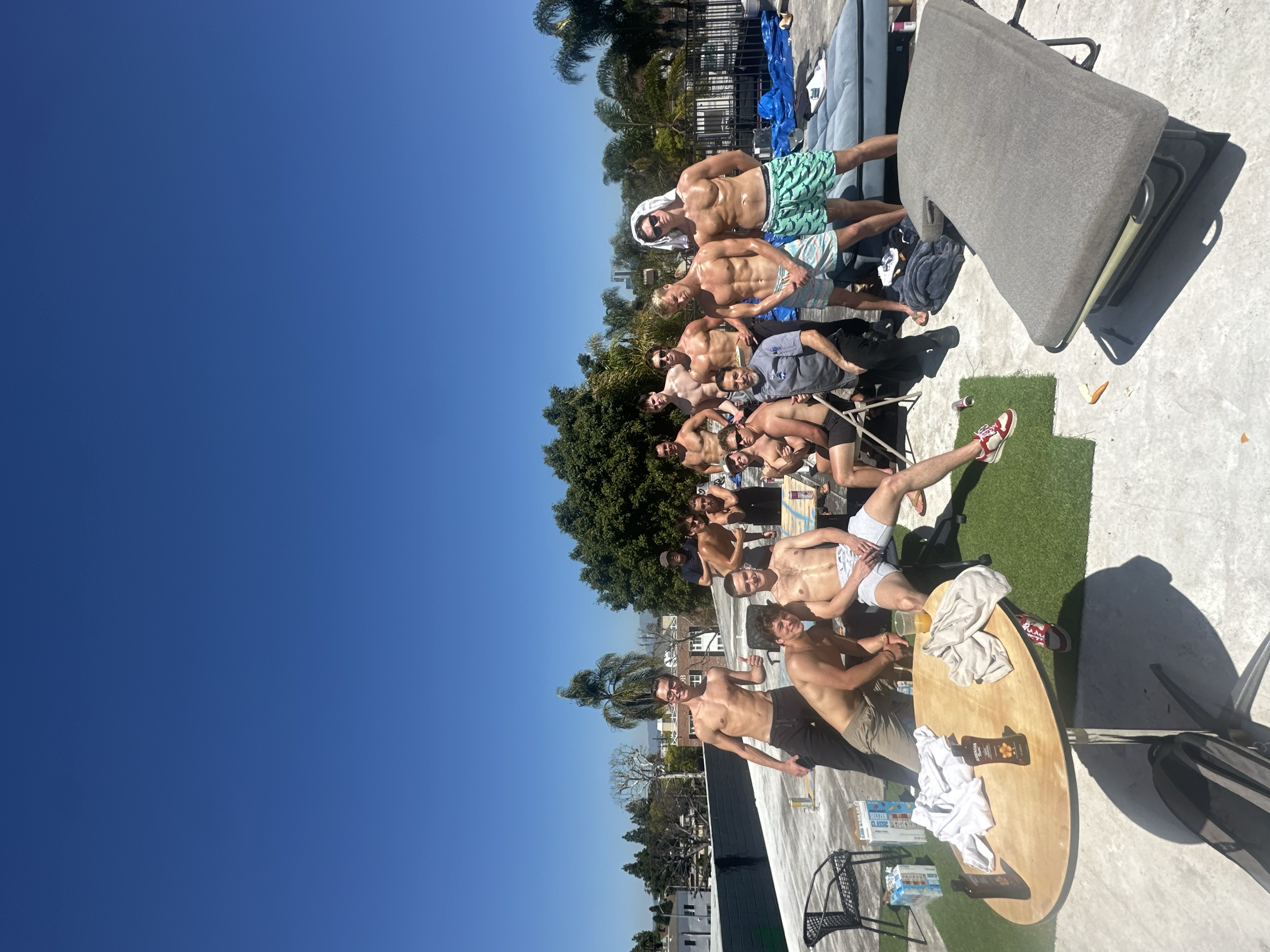 A group of people in swimwear gathered together outdoors on a sunny day, posing for a photo on a concrete platform near a pool, with trees and a clear blue sky in the background.