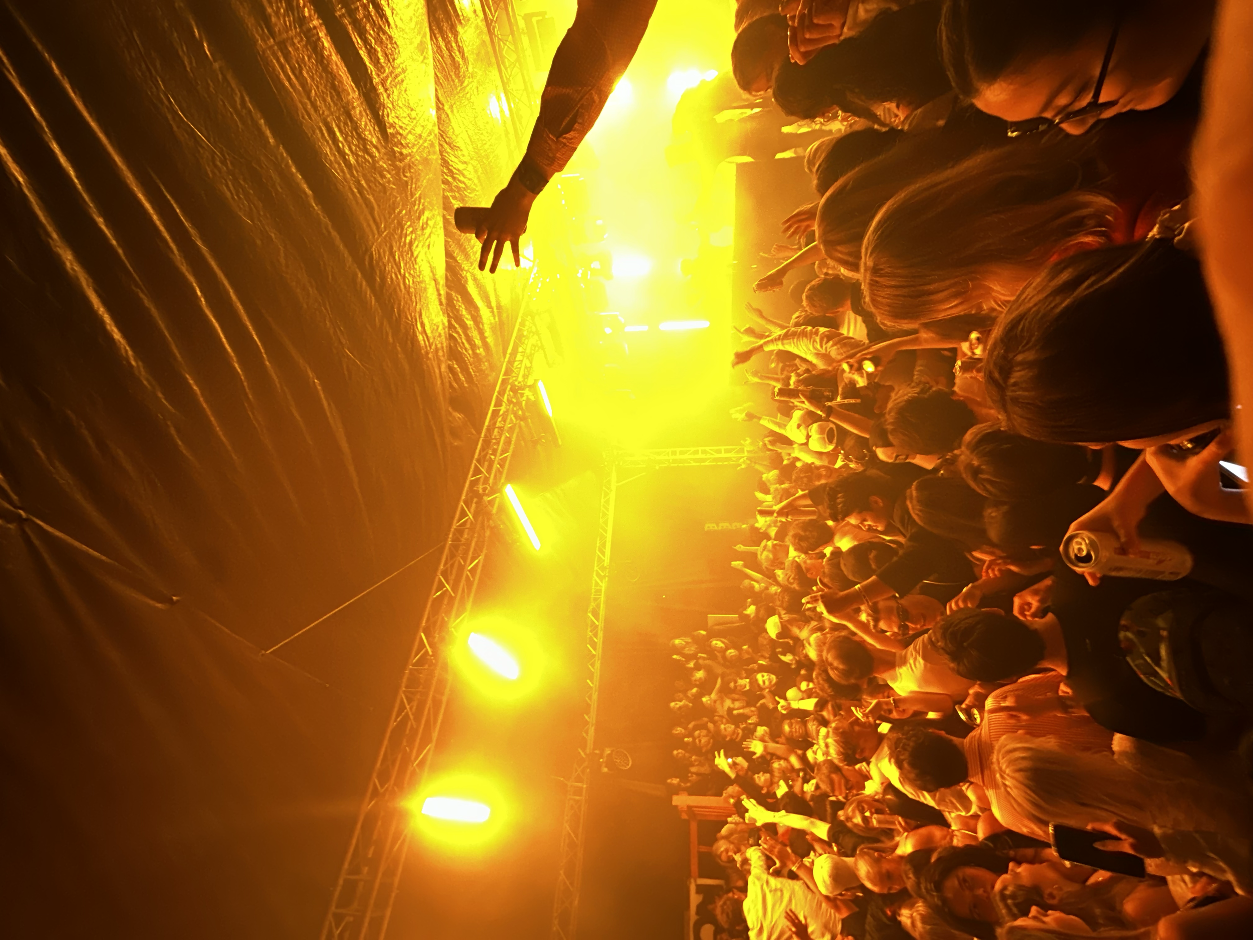 A large crowd of people in a concert or nightclub, with bright yellow and orange stage lights illuminating the scene. The audience is standing close together, some with drinks, enjoying the live performance.