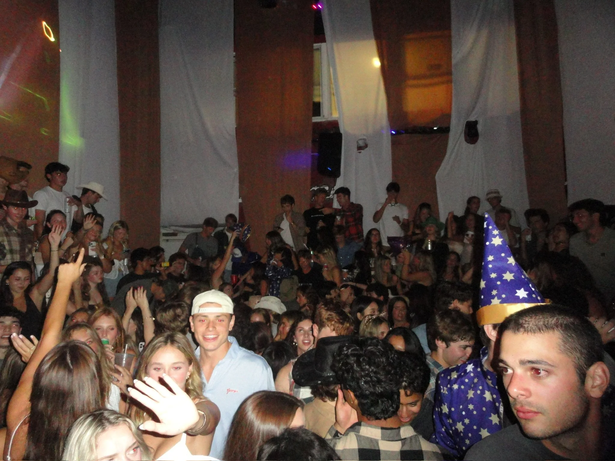 A crowded party scene with young people, some wearing party hats, dancing and socializing indoors.