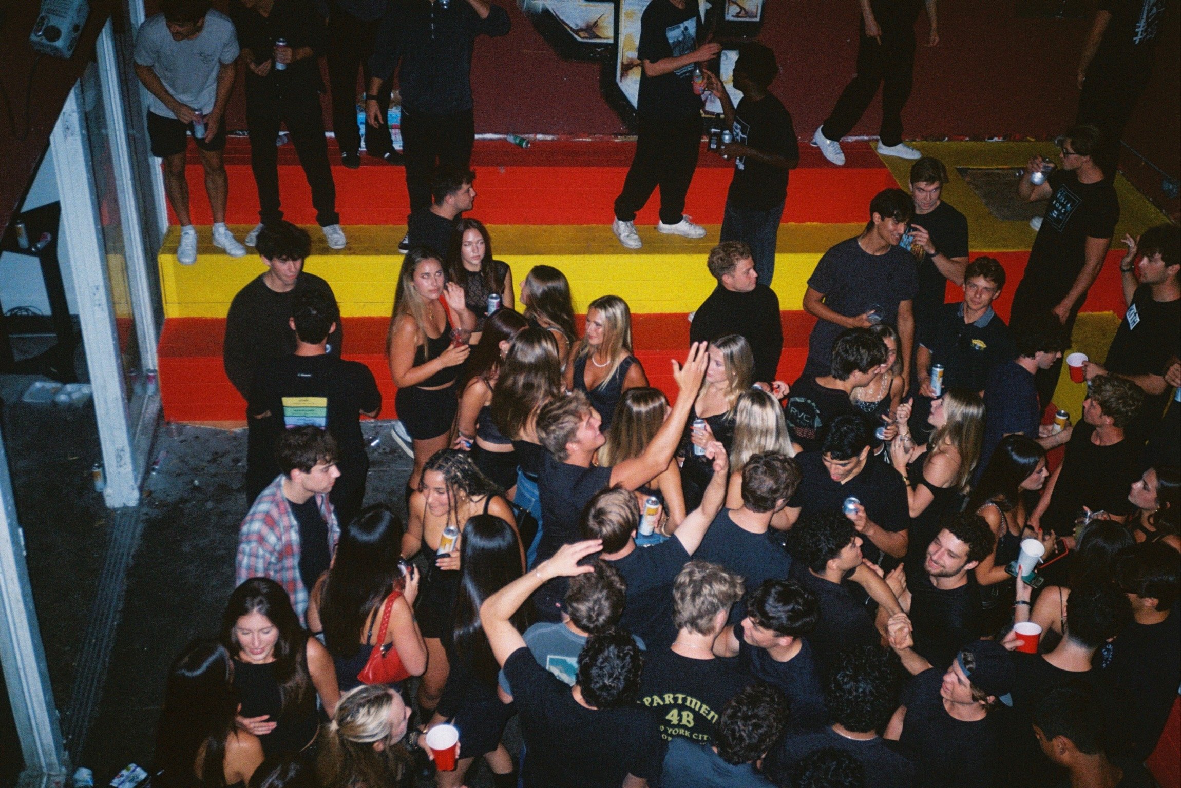 Crowd of young people at a party, dancing and socializing, with some holding drinks, on a staircase with colorful steps