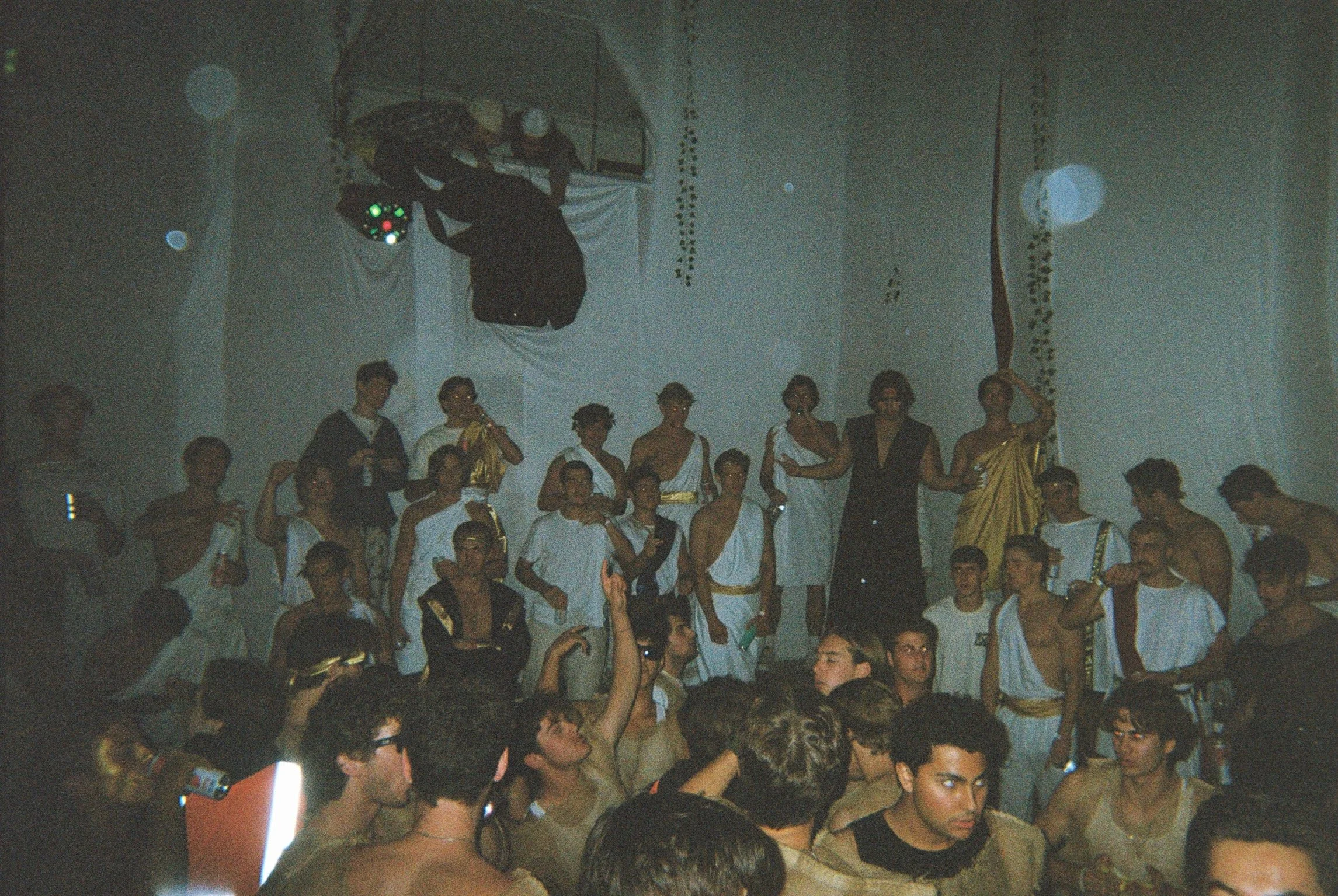 People dressed in ancient Greek or Roman costumes celebrating at a party or event, with some standing on a stage or platform and others sitting or dancing in front.