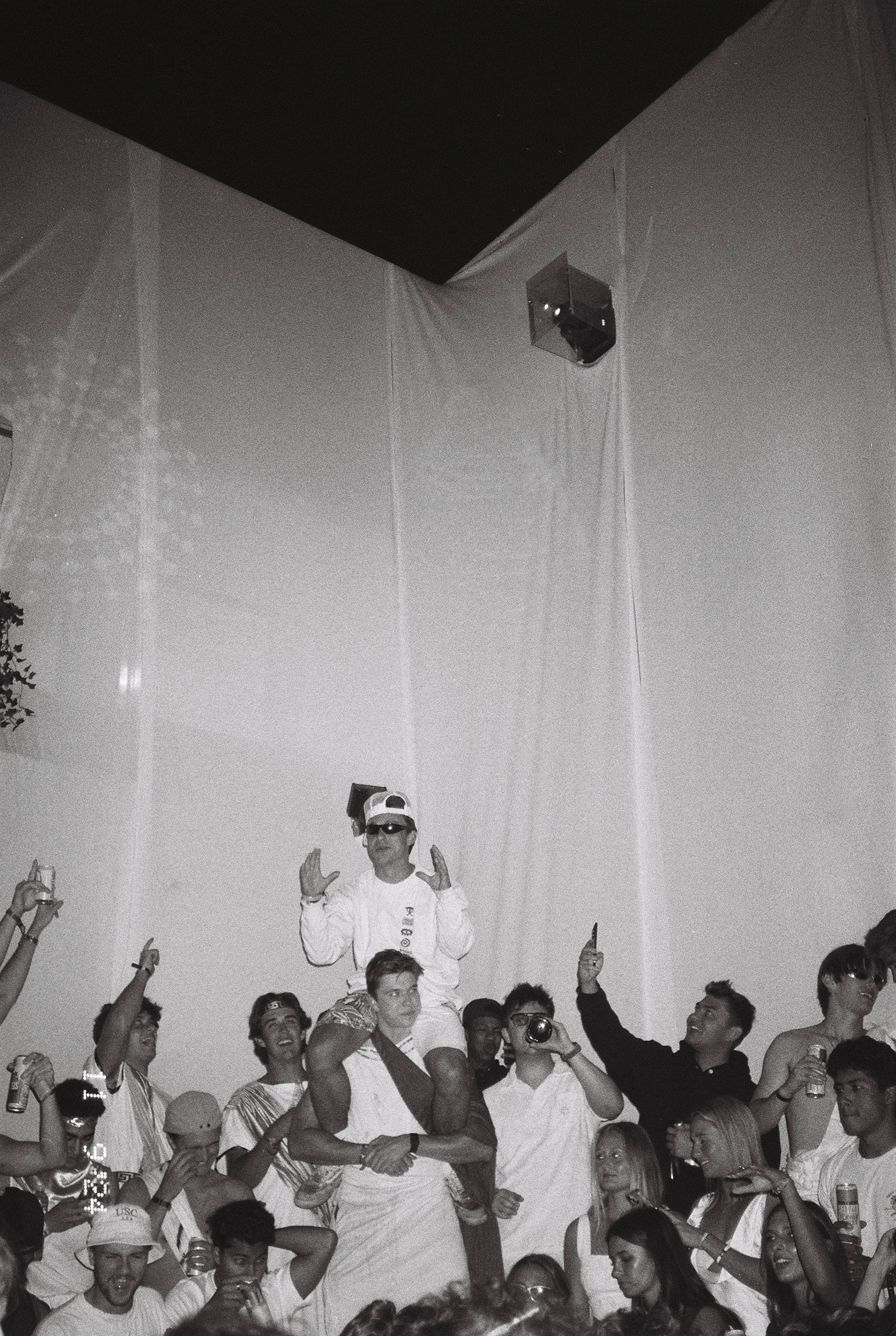 A group of young people at a party, some holding drinks, with one person being lifted on someone’s shoulders, wearing sunglasses and a cap, surrounded by others taking photos and enjoying the event.