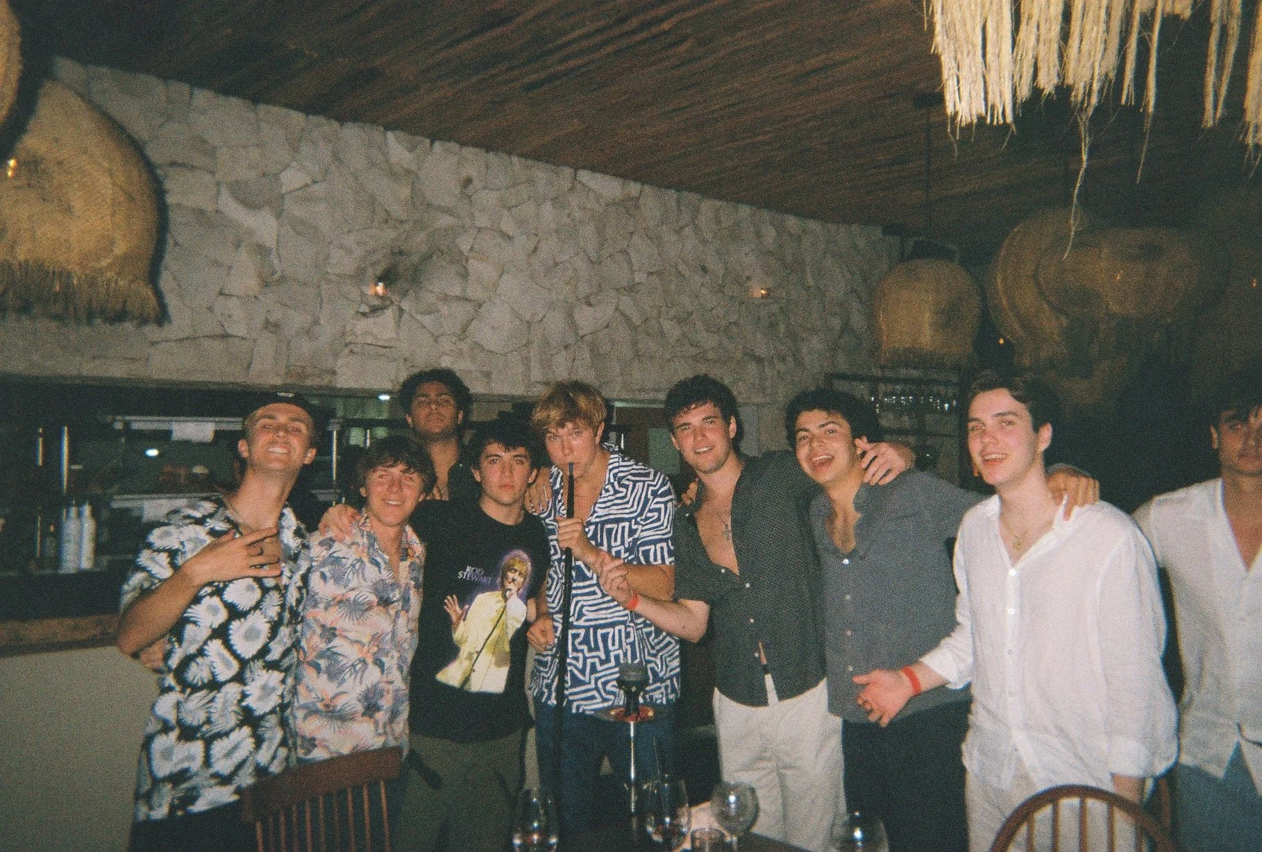 Group of young men smiling and posing together in a restaurant or bar setting, some with arms around each other, with a stone wall background and hanging baskets.