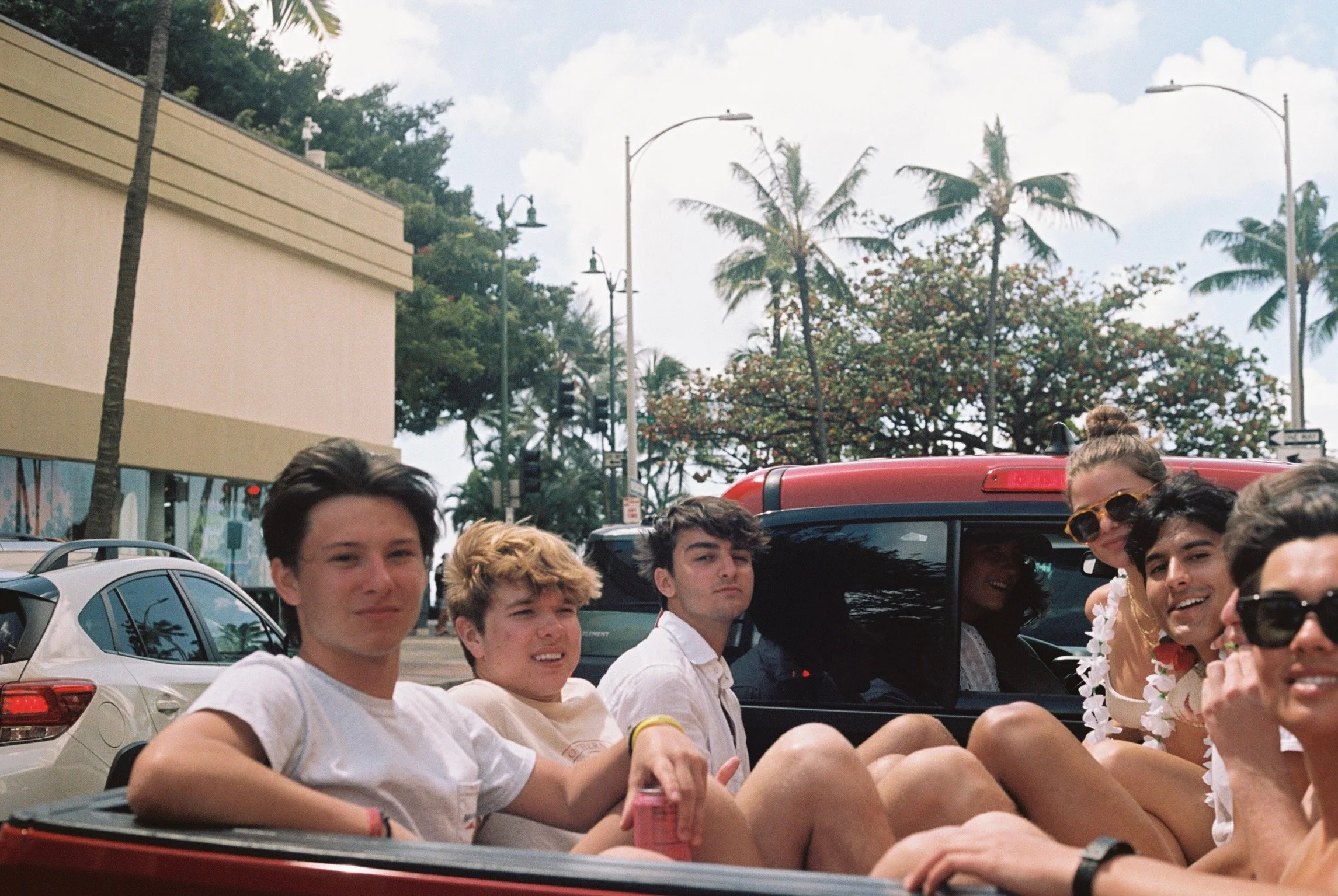 A group of young people sitting in the back of a red pickup truck on a sunny day, with palm trees and a building in the background.