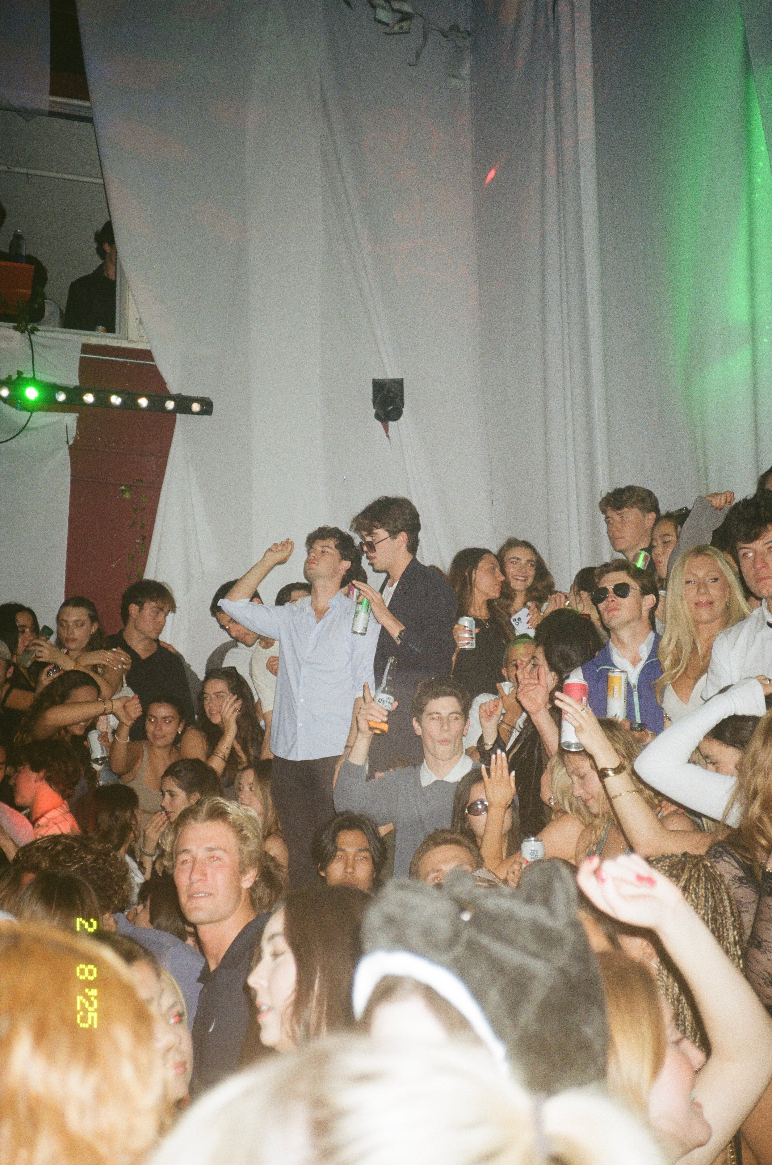 Crowd of young people at a party or nightclub, dancing and holding drinks, with some wearing sunglasses and others raising their hands in the air.