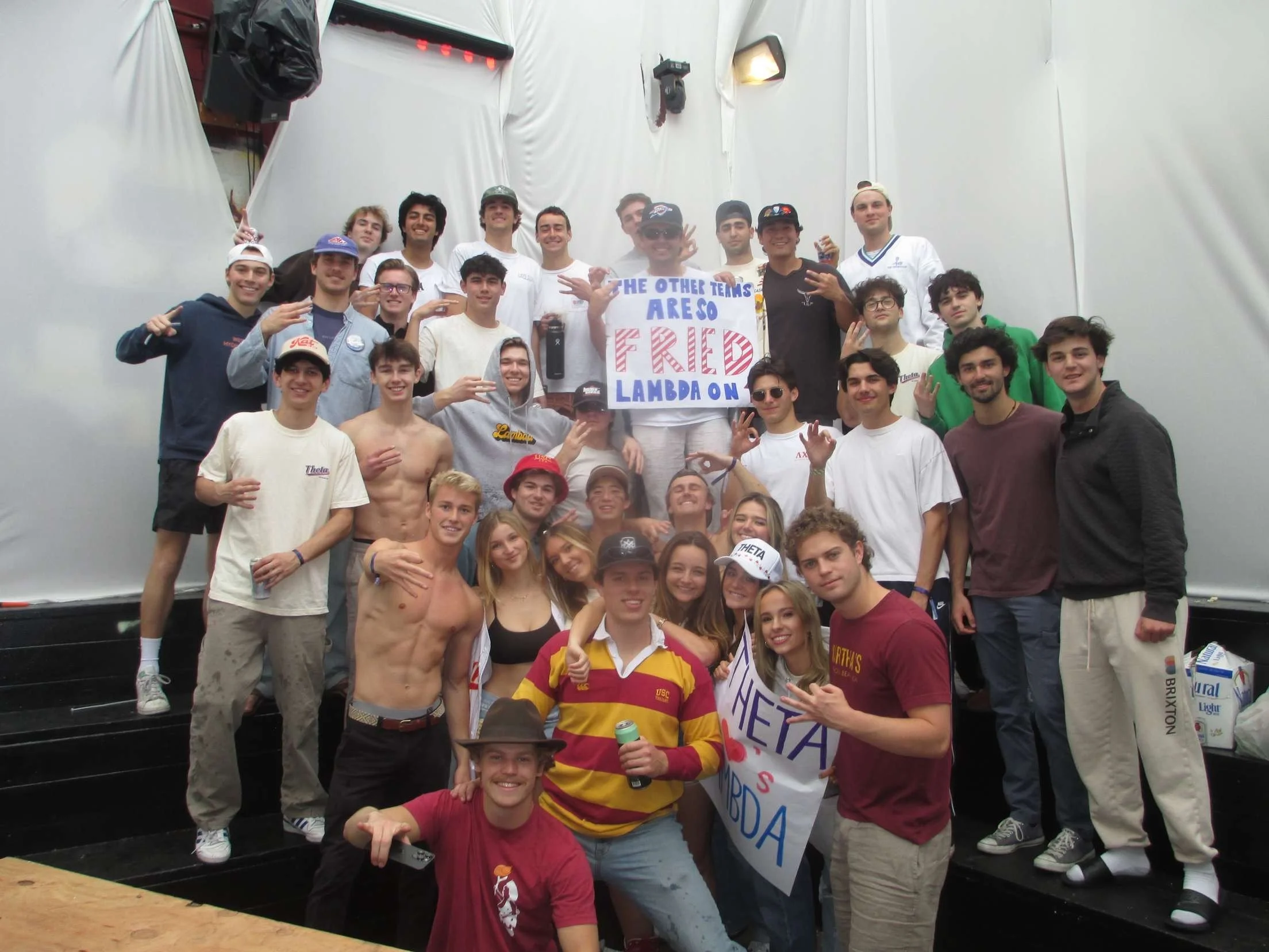 Group of young adults and college students celebrating indoors, holding signs and making hand gestures, group photo on a small stage with white walls and screens.