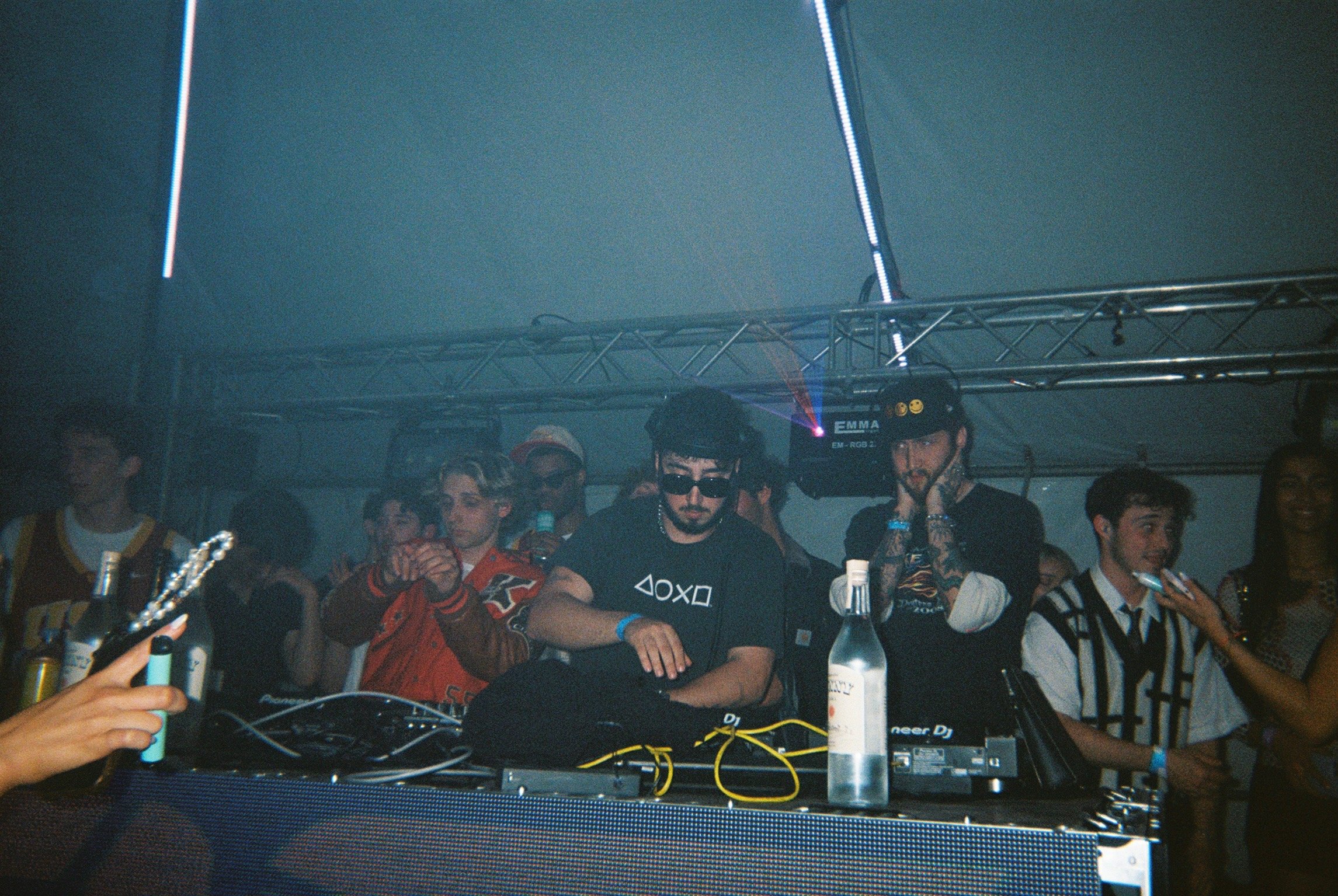 A DJ with sunglasses and headphones stands behind a mixing table at a party, surrounded by a crowd of young people dancing and listening, with party lighting in a tent.