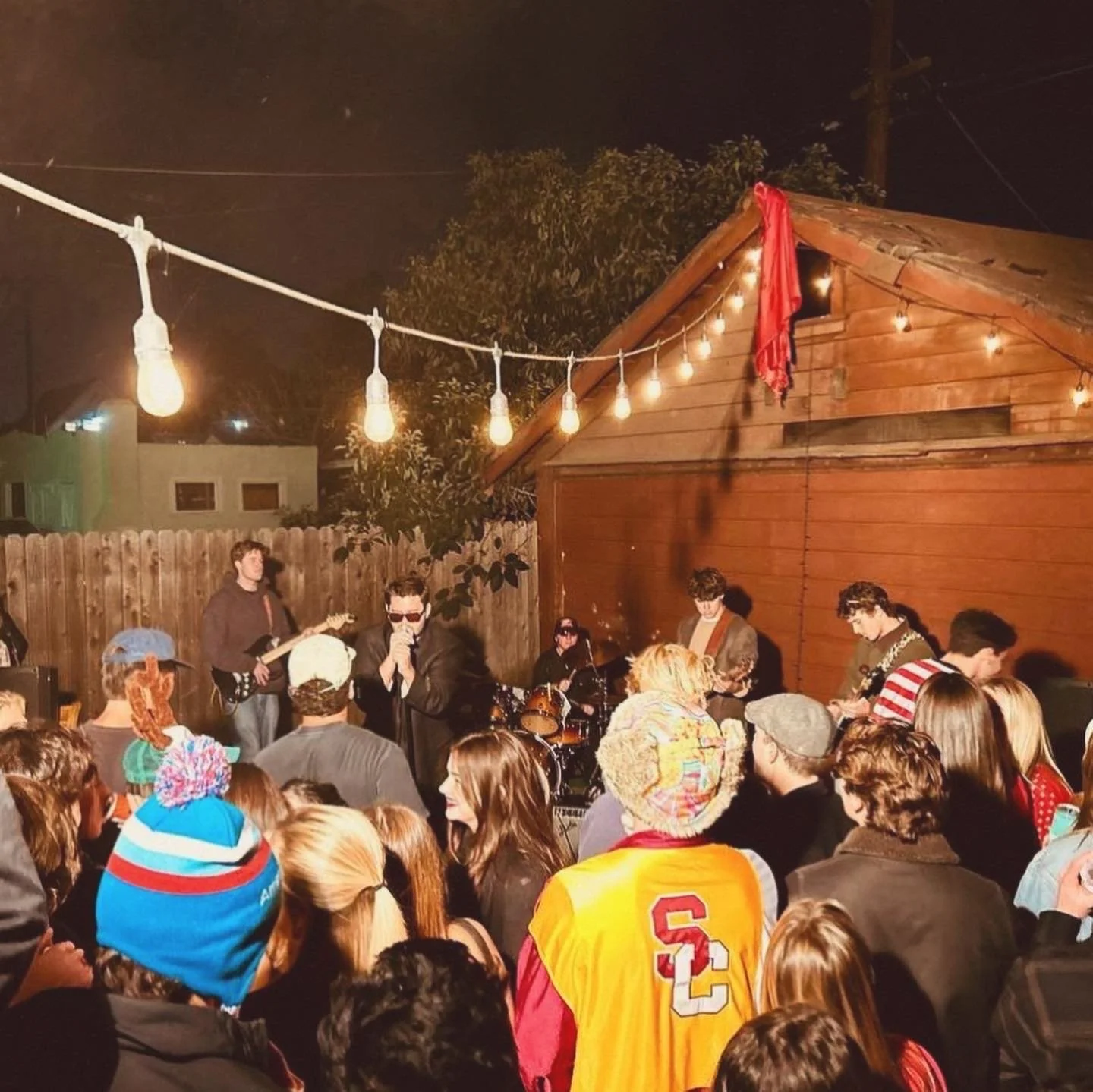 People gathered outdoors listening to a live band perform at night, with string lights hanging overhead.