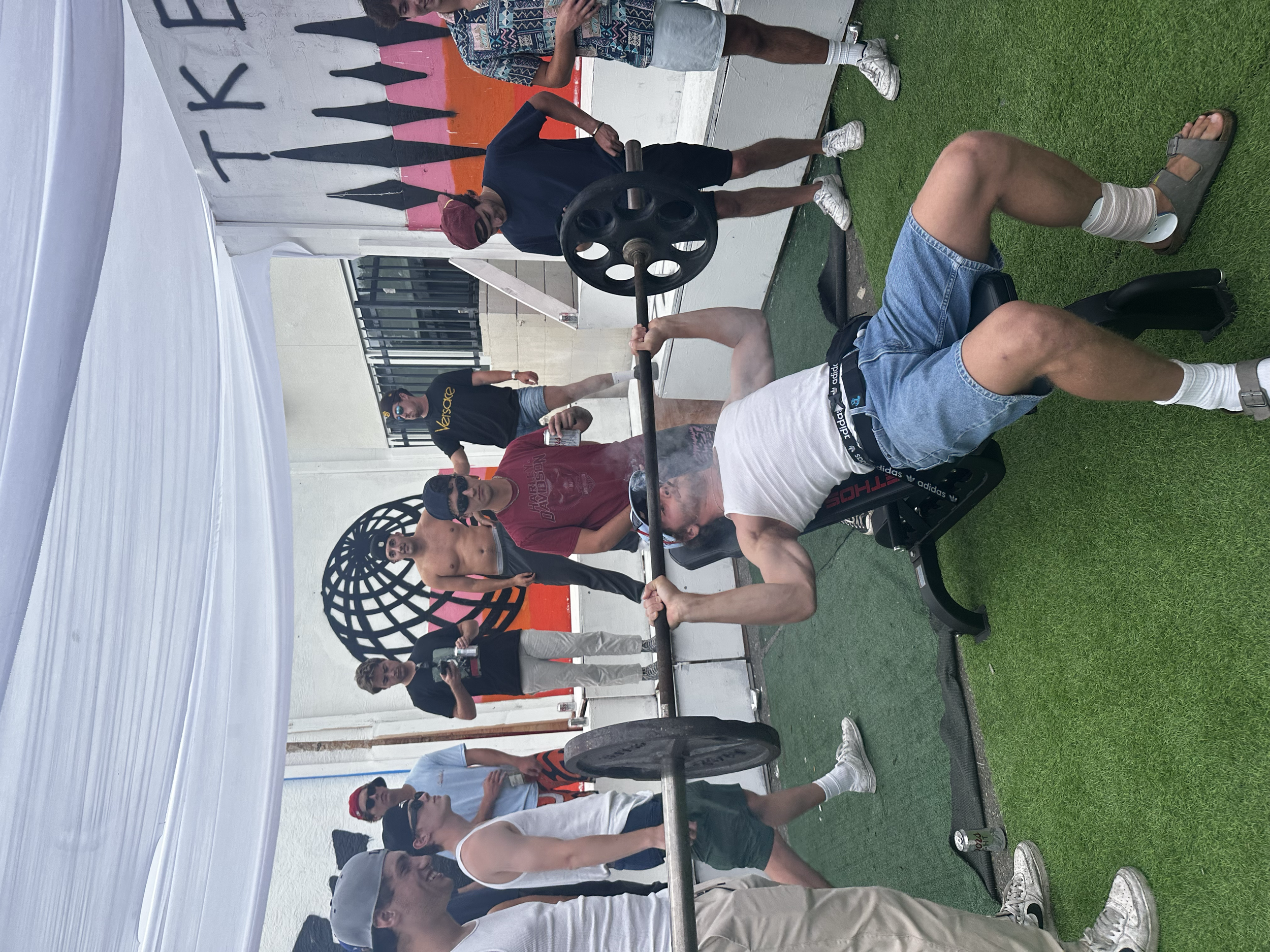 A man performing a flat bench press exercise with a barbell at an outdoor gym. Several people watch, some taking photos. The background features a mural with the word 'TIK' and an abstract globe design.
