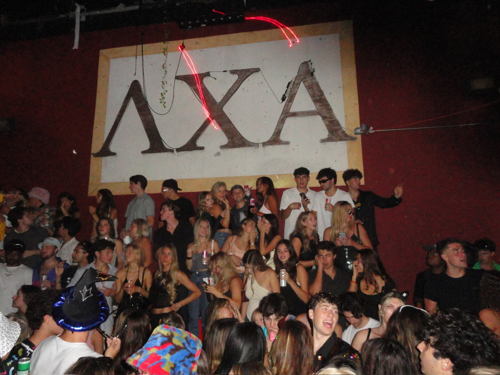 Crowd of young people at party with large banner reading 'XXA' in the background.