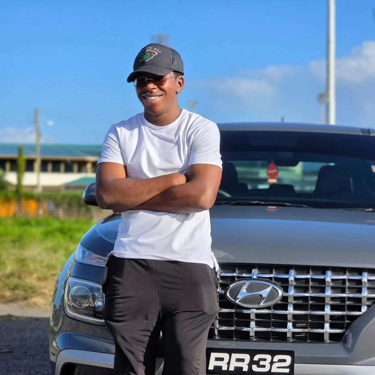 A young man wearing a cap, sunglasses, a white t-shirt, and black shorts standing with arms crossed in front of a gray Hyundai car with a "RR 32" license plate, outdoors on a sunny day with a blue sky.
