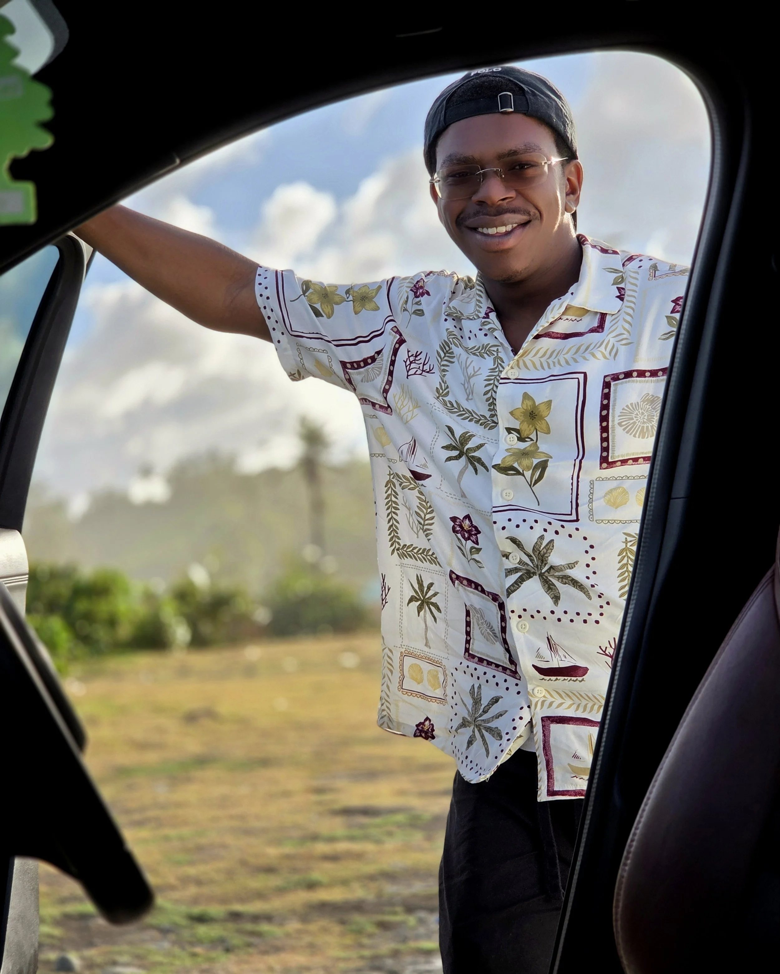A smiling man wearing sunglasses, a black cap, and a floral patterned shirt, leaning into the open doorway of a vehicle with a landscape of grass and trees in the background.