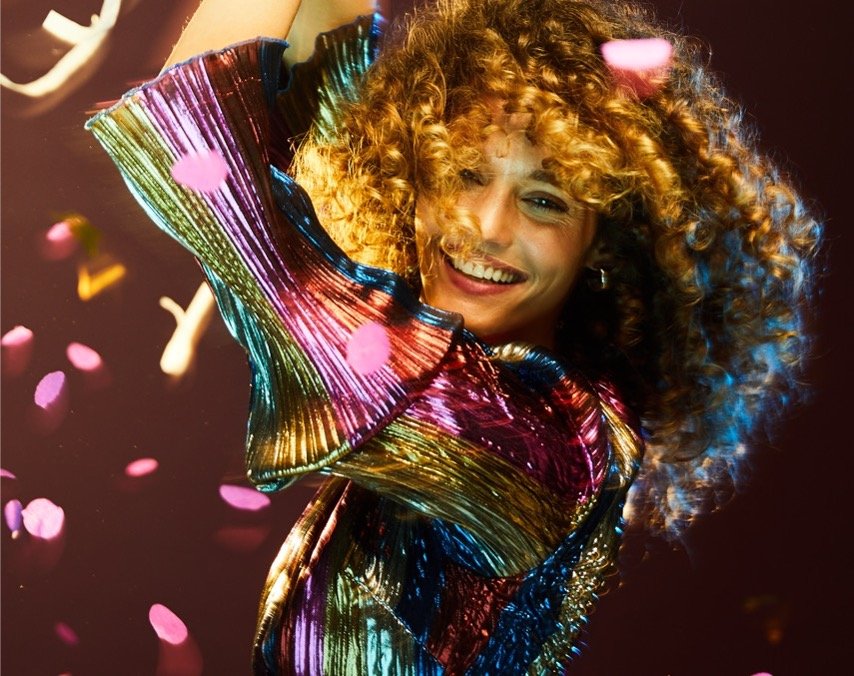 Woman with curly hair smiling, wearing a colorful shiny dress, surrounded by falling pink confetti.