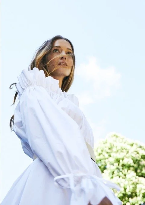 A woman in a white ruffled dress standing outdoors with a blue sky and some greenery in the background.