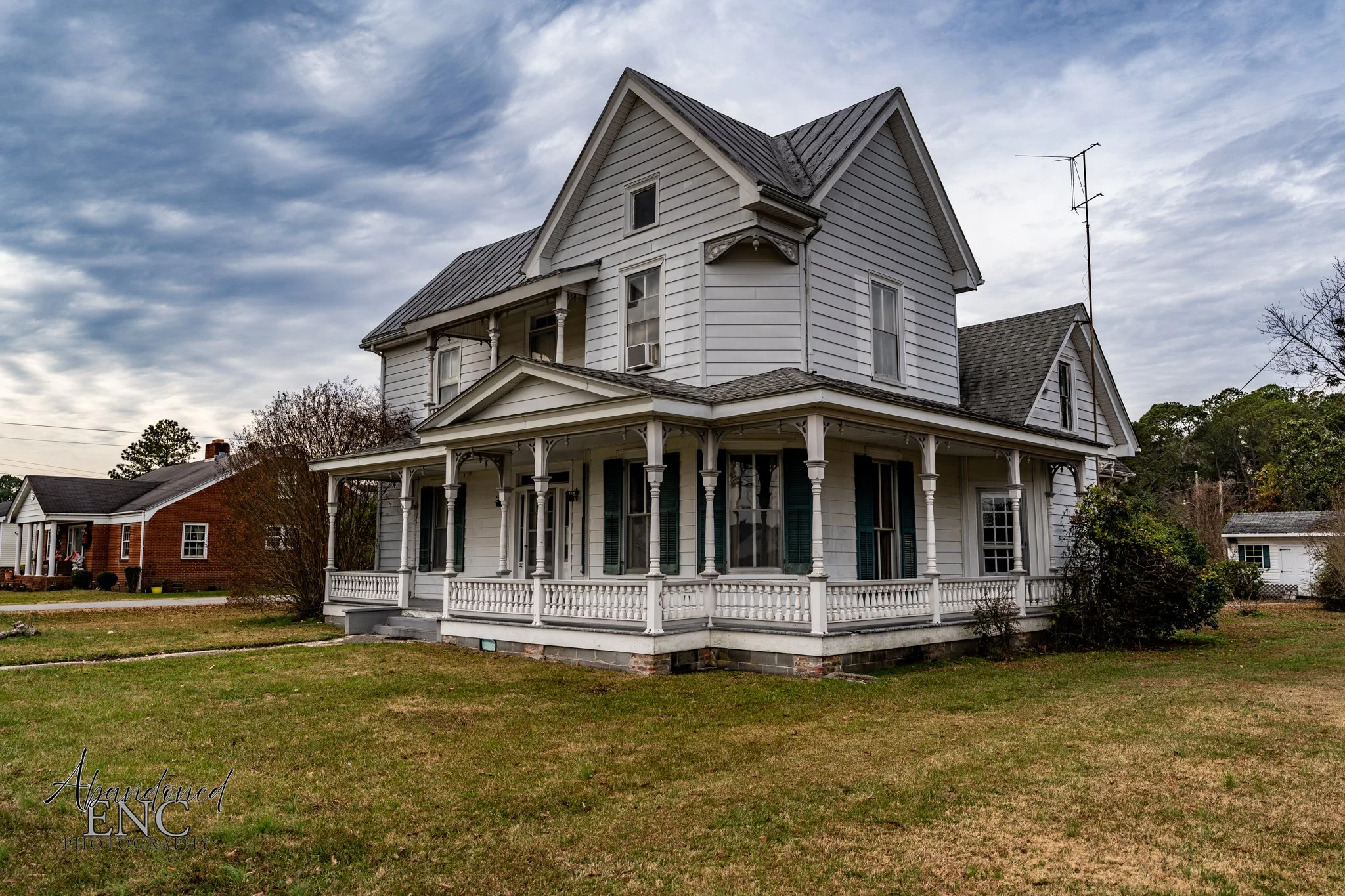 A large, two-story American house with white wooden siding and a wrap-around porch with decorative railings, green shutters, and multiple windows, under a cloudy sky.