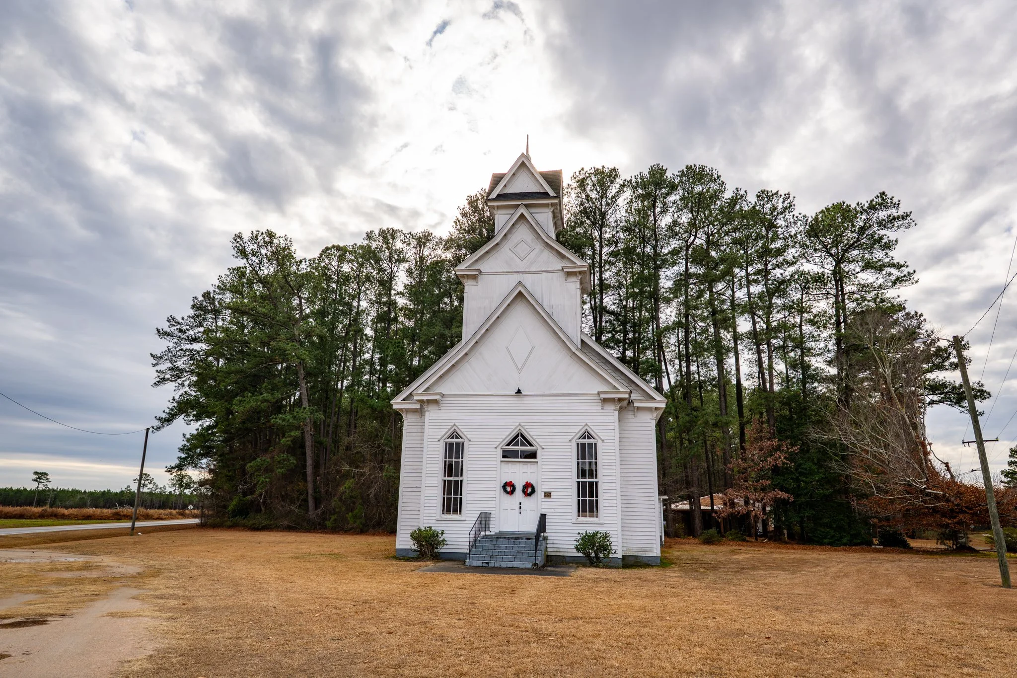 A white wooden church with a steeple, decorated with holiday wreaths on the front door, surrounded by trees and a cloudy sky.