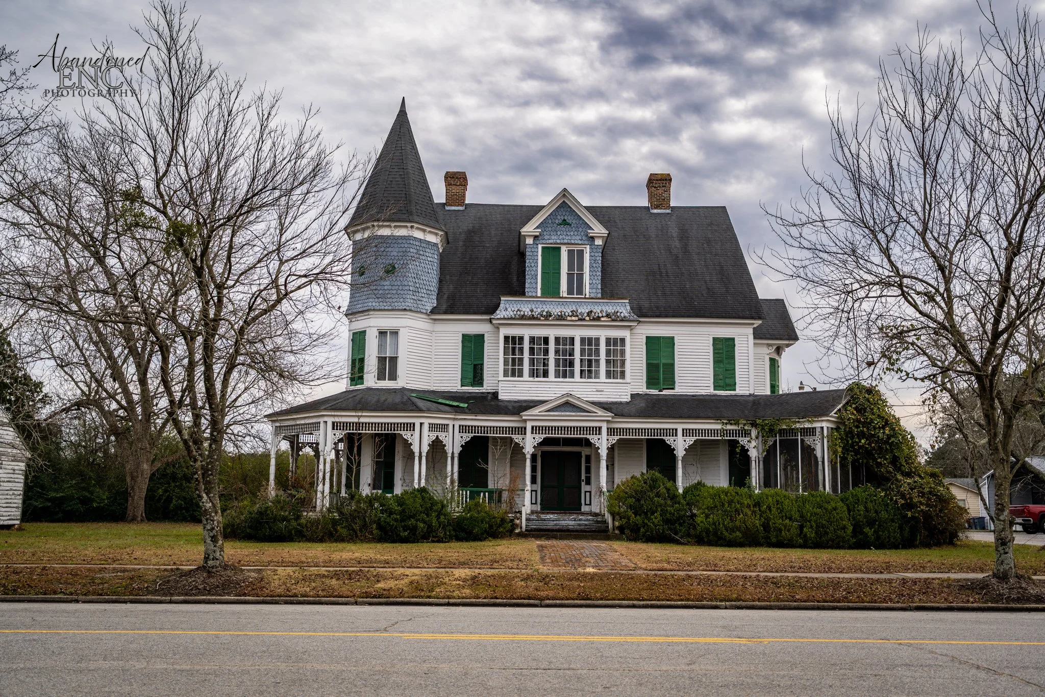 A large, Victorian-style house with white siding, green shutters, and a steeply pitched roof, situated behind a grassy lawn and a street.