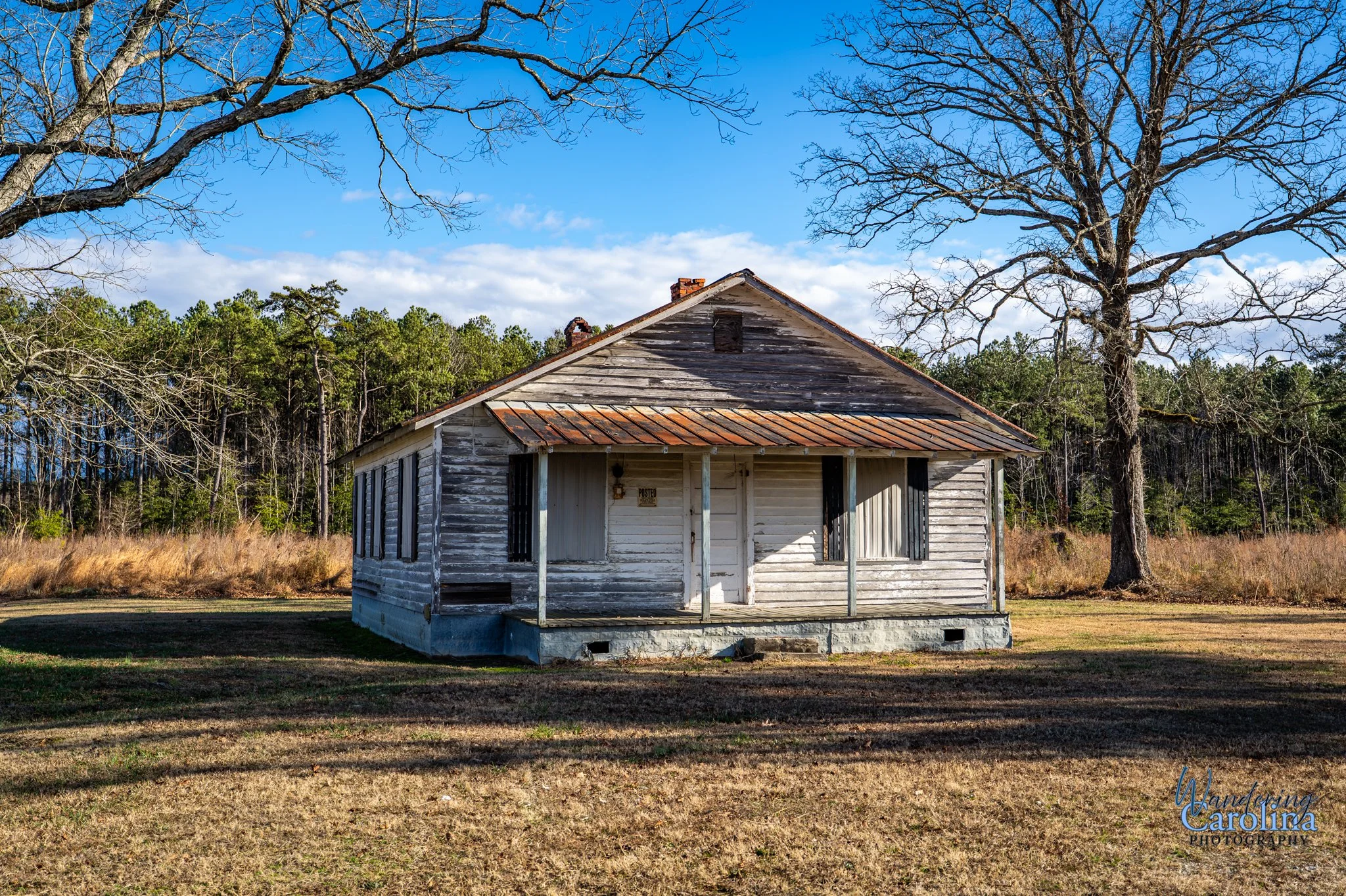 An old, weathered wooden house with a rusted metal roof in a rural area, surrounded by grass and large leafless trees with a backdrop of forested trees and a partly cloudy sky.