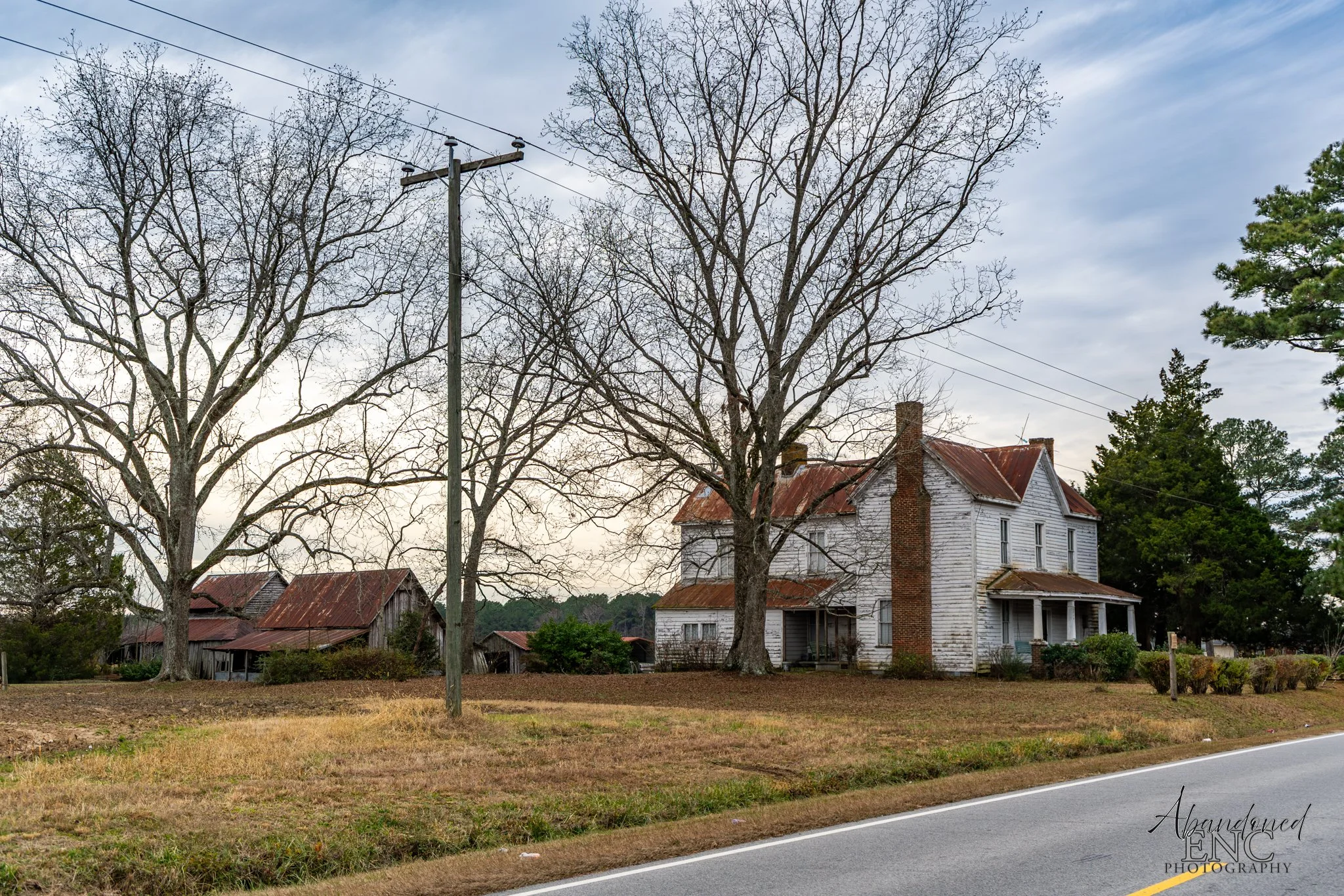 An old, abandoned white house with a rusty roof and chimney, surrounded by leafless trees, with a clear sky in the background and a paved road in the foreground.