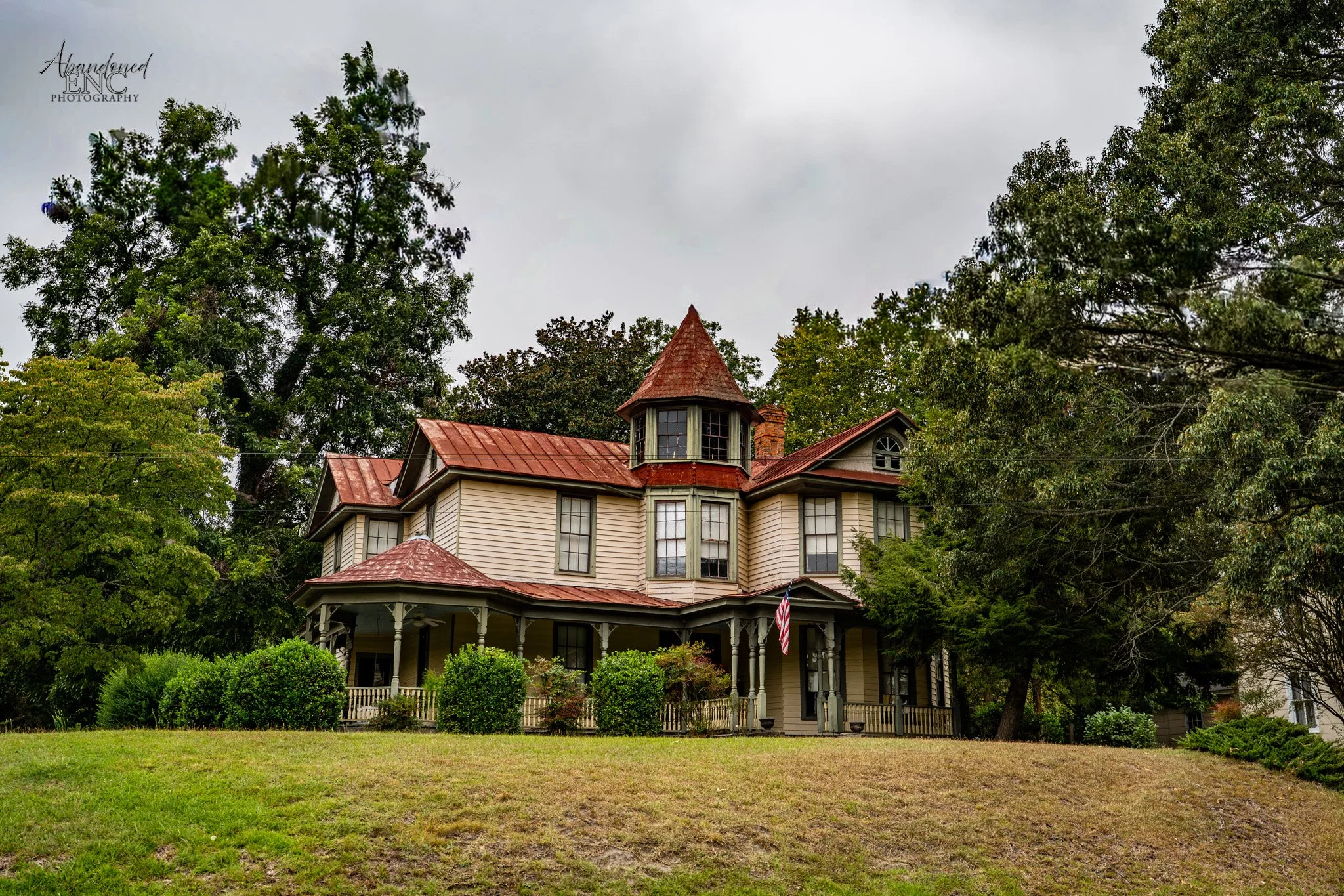 A large Victorian-style house with beige siding, red metal roof, and a pointed turret, surrounded by green trees and bushes, with a lawn in the front.