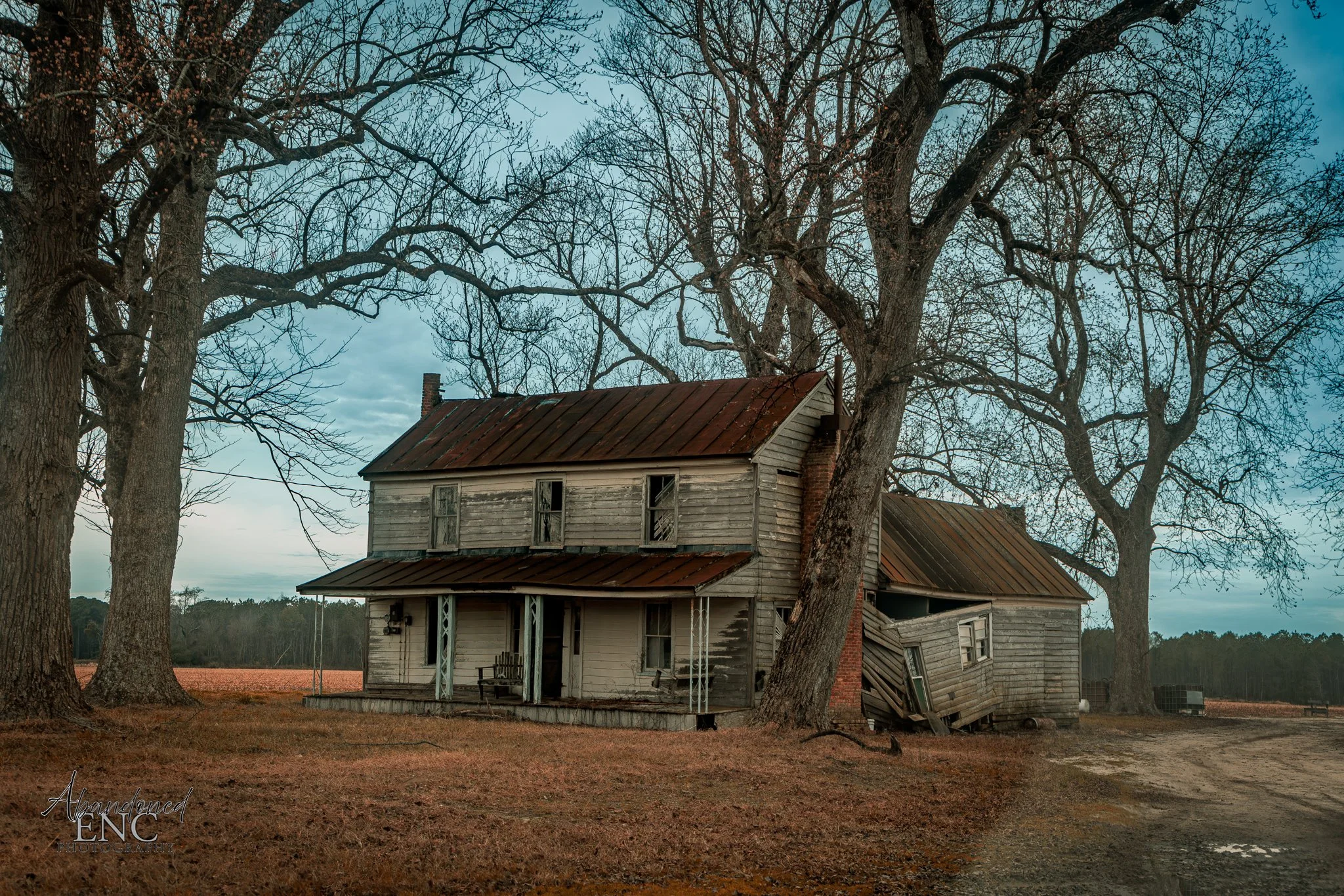 An old, abandoned two-story wooden house with a rusted metal roof, featuring boarded windows, and leaning to one side. It is surrounded by leafless trees, with a dirt path nearby, under a cloudy sky.