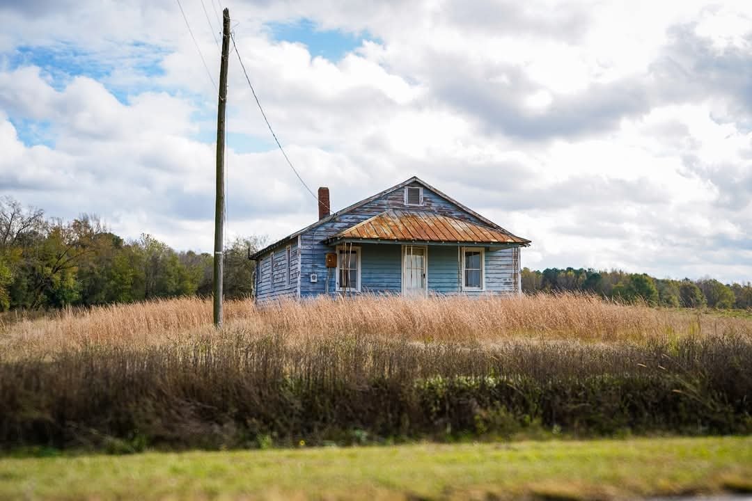 An old, weathered blue house with a rusted metal roof sits in a field of tall grass, under a partly cloudy sky.