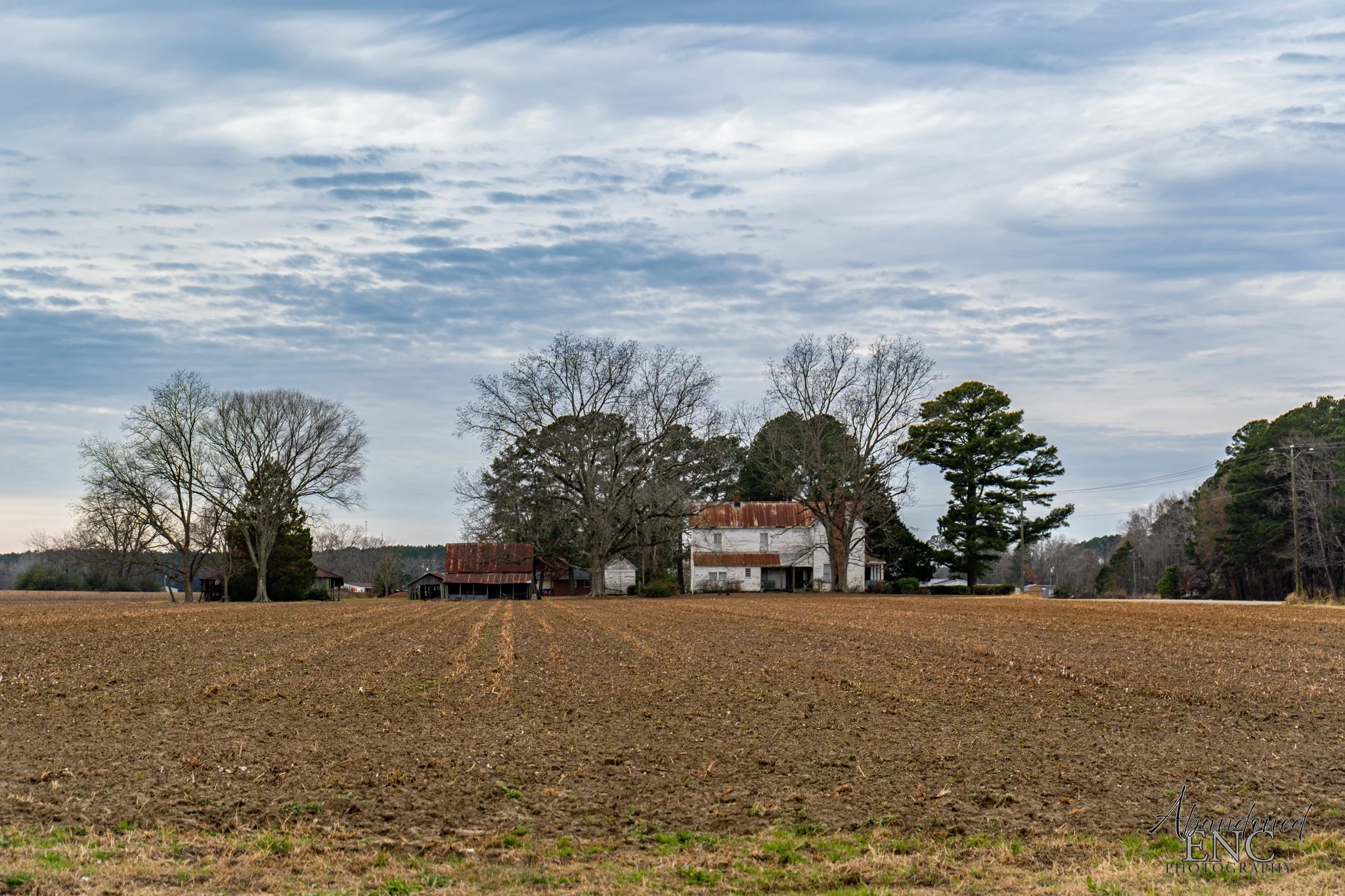 A rural landscape with an overcast sky, a plowed field in the foreground, and an old farmhouse with trees surrounding it in the background.