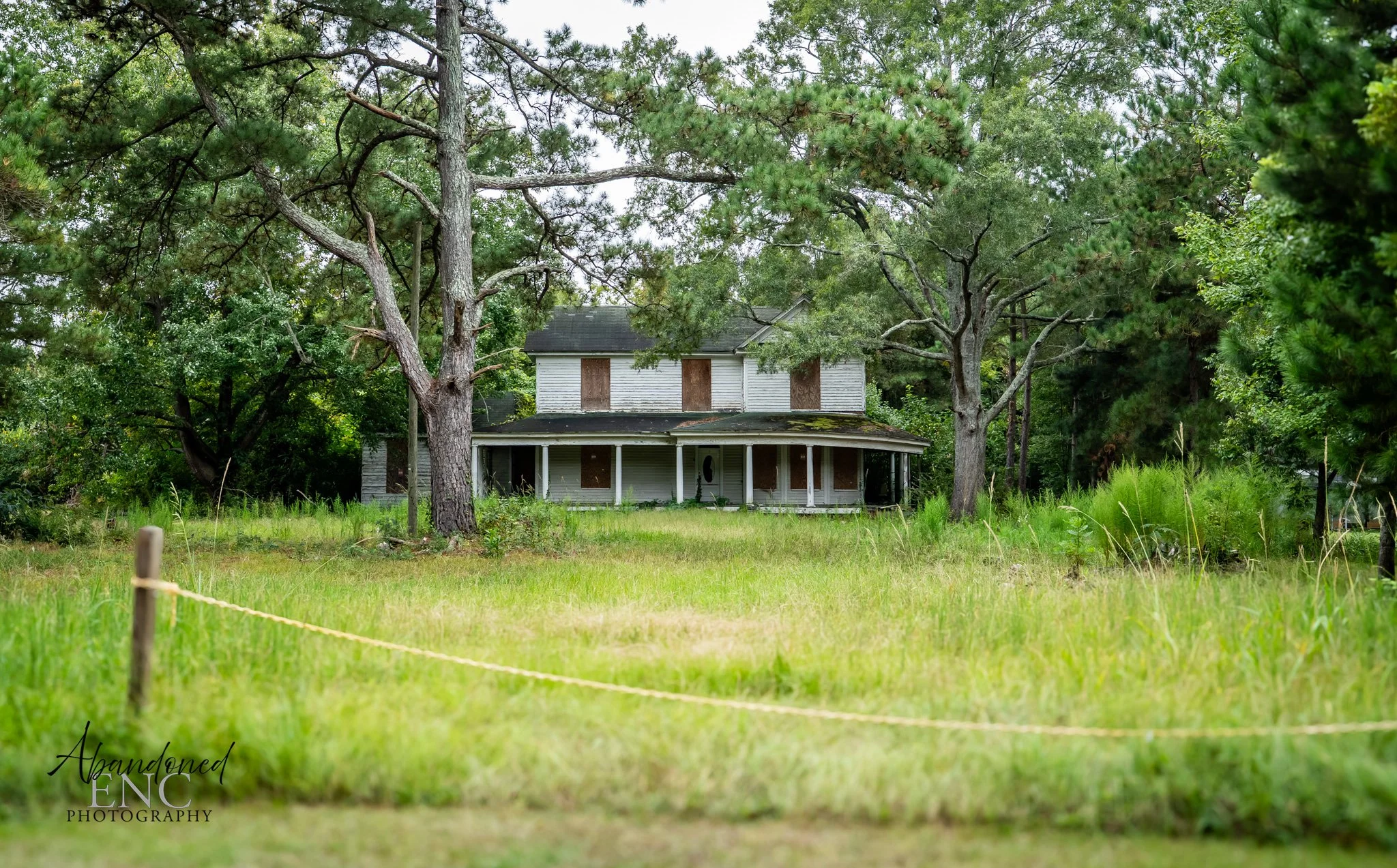 An abandoned, two-story house with boarded-up windows, surrounded by large trees and overgrown grass.