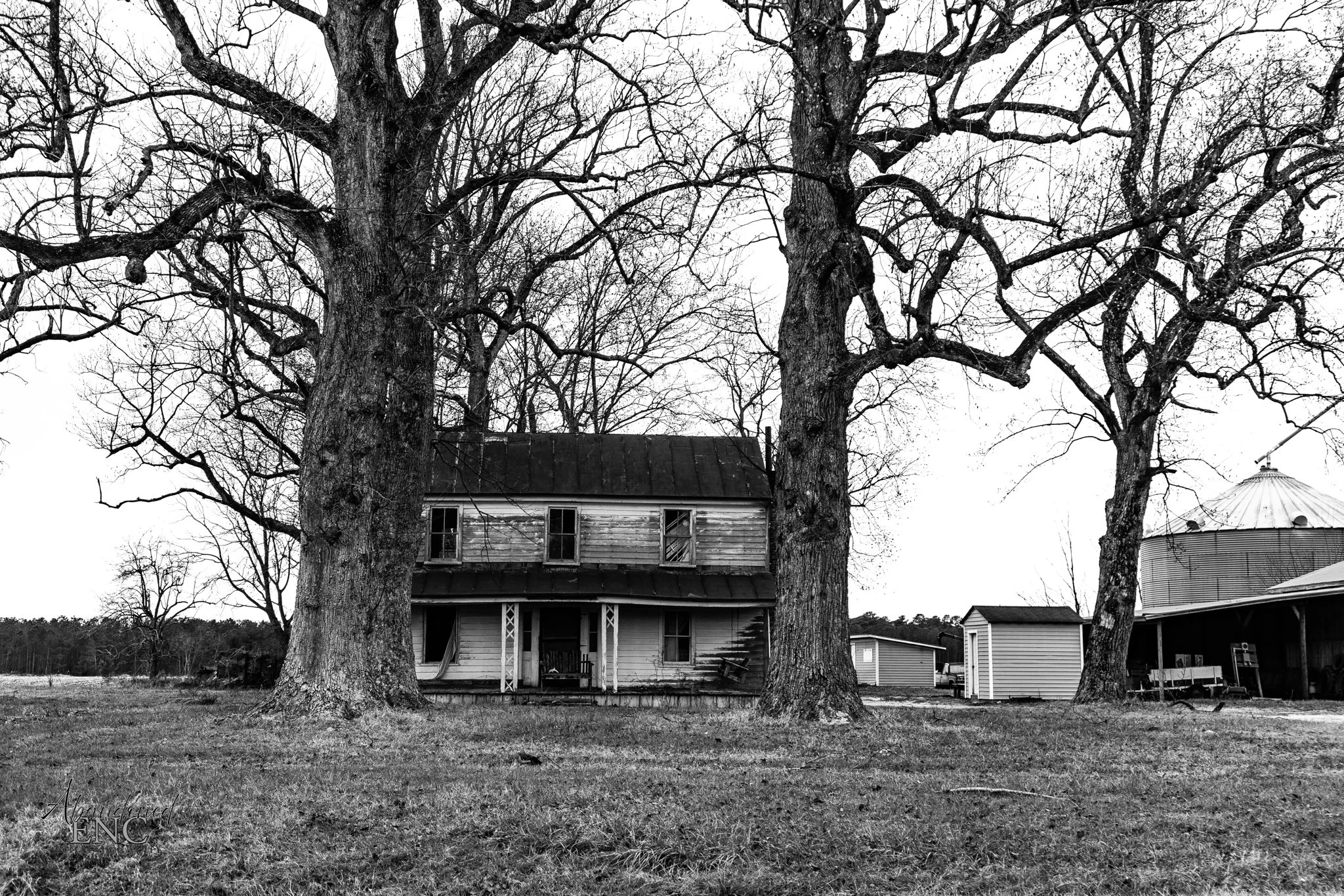 Black and white image of an old, two-story house with large, leafless trees in the front yard, and a barn and storage shed in the background.