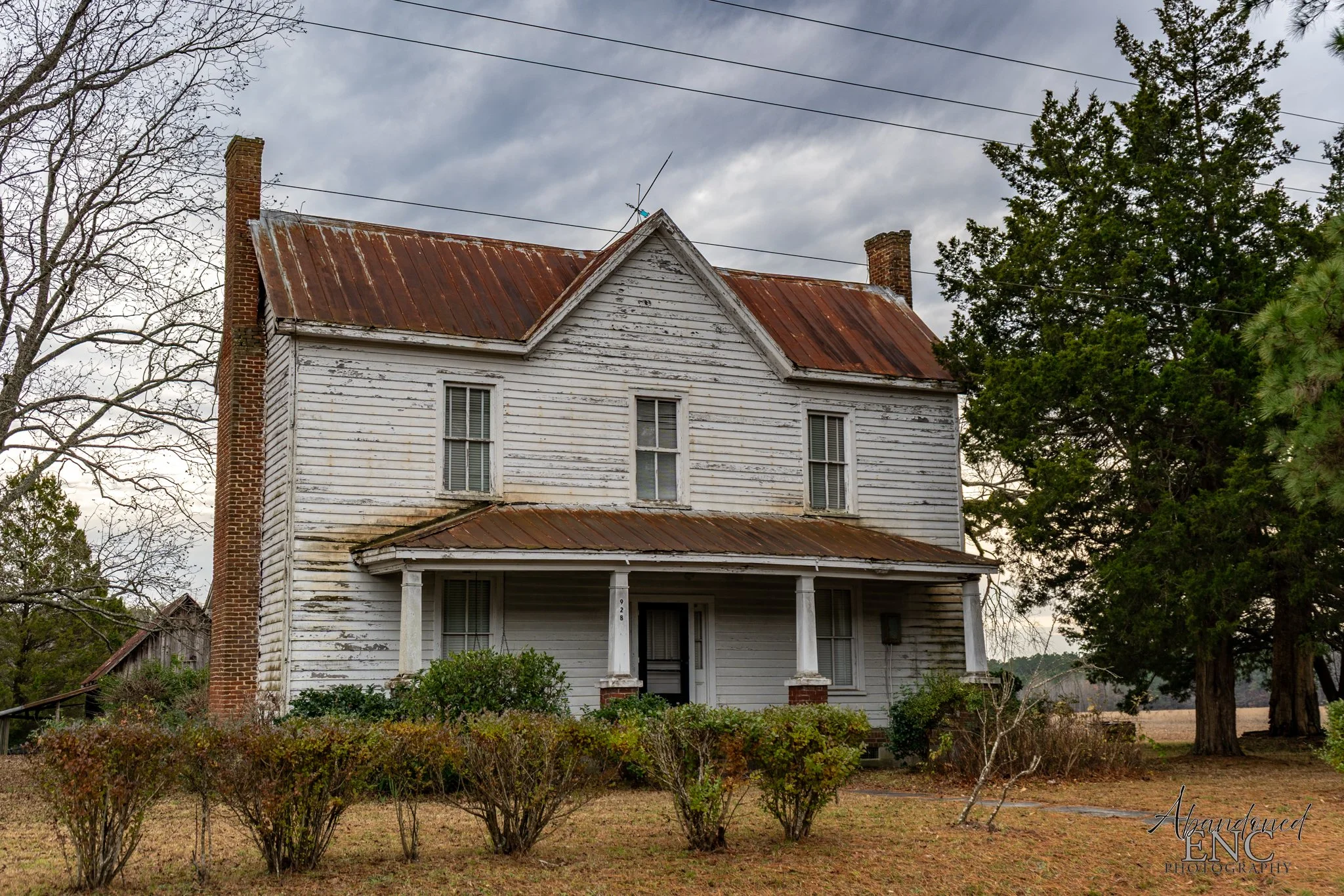 An old, two-story, white wooden house with a rusty metal roof and chimney, surrounded by bushes and trees under a cloudy sky.