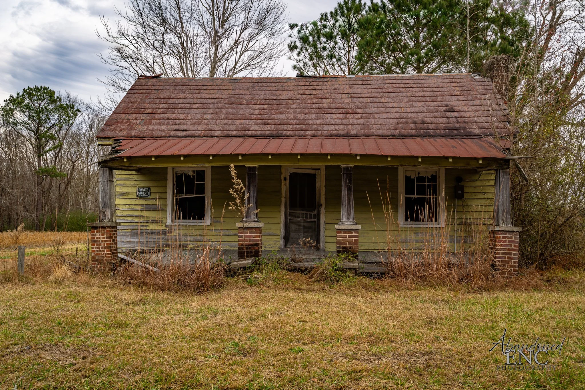 An abandoned, dilapidated house with green paint, broken windows, and a sagging porch, surrounded by overgrown grass and leafless trees.