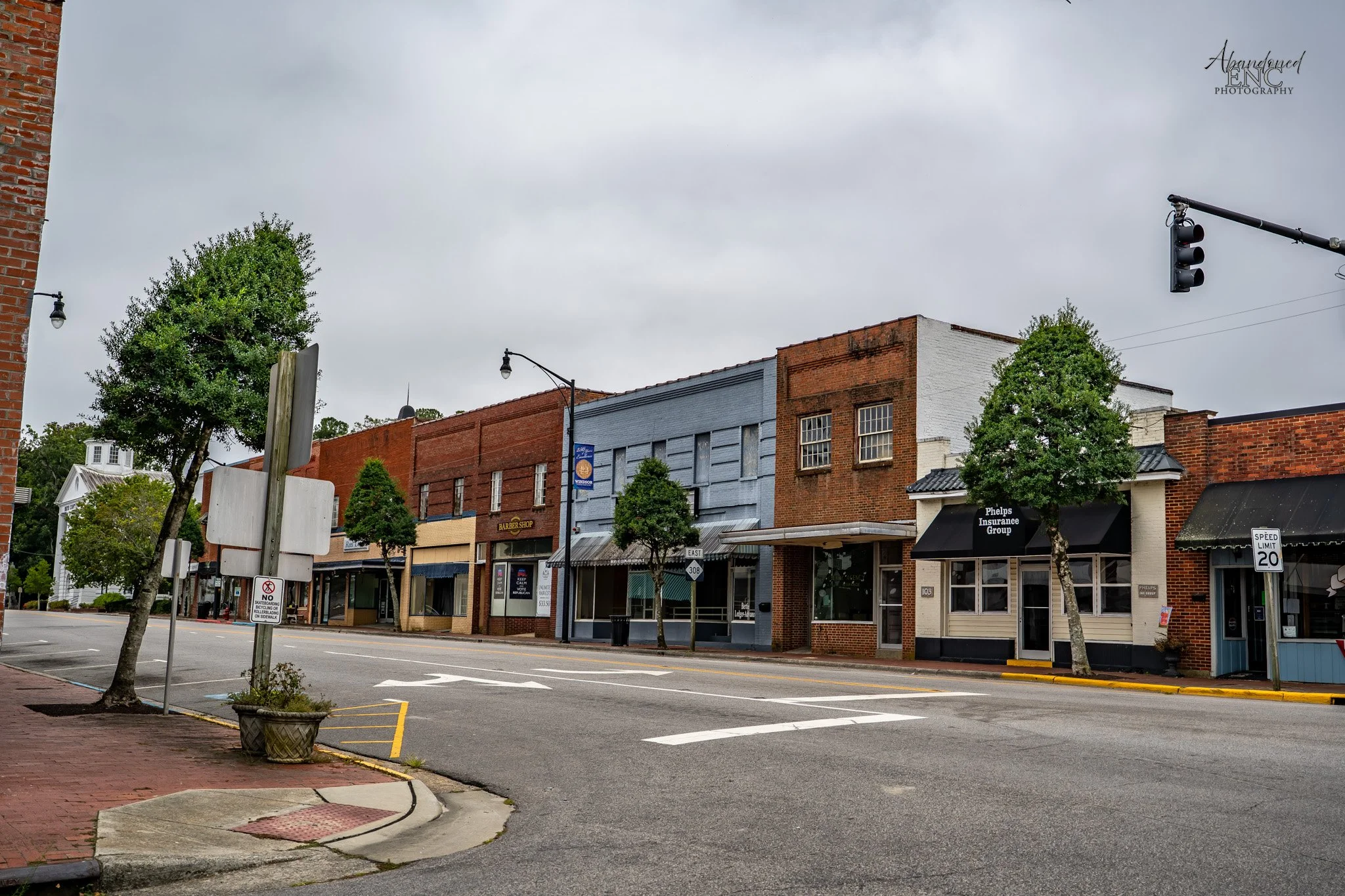Empty street in a small town with brick and wooden storefronts, some trees, street signs, traffic light, and overcast sky.