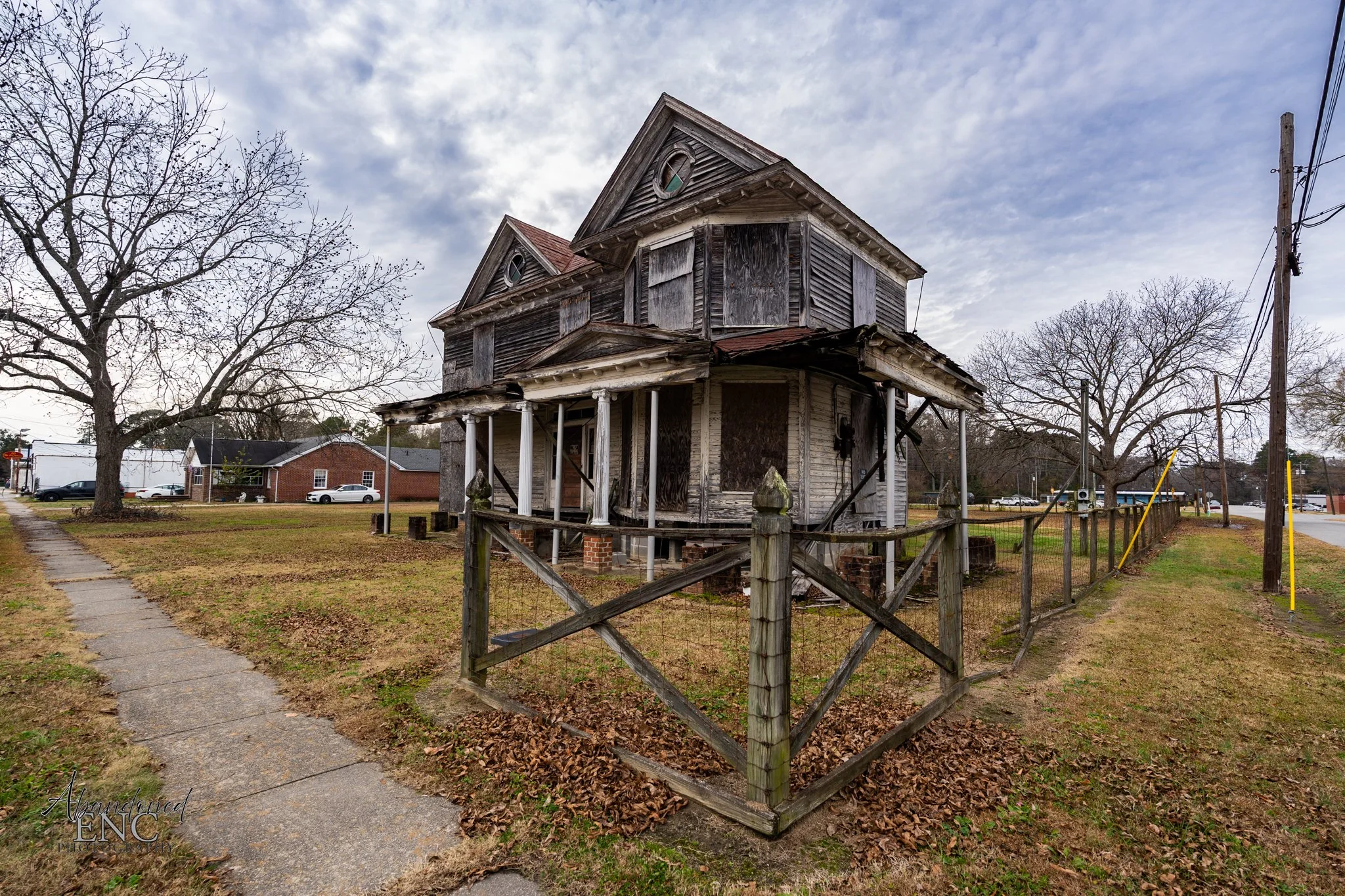 An old, abandoned, two-story wooden house with a porch, boarded-up windows, and a deteriorated exterior, surrounded by a wooden fence and laying in a neighborhood with trees, cars, and power lines.