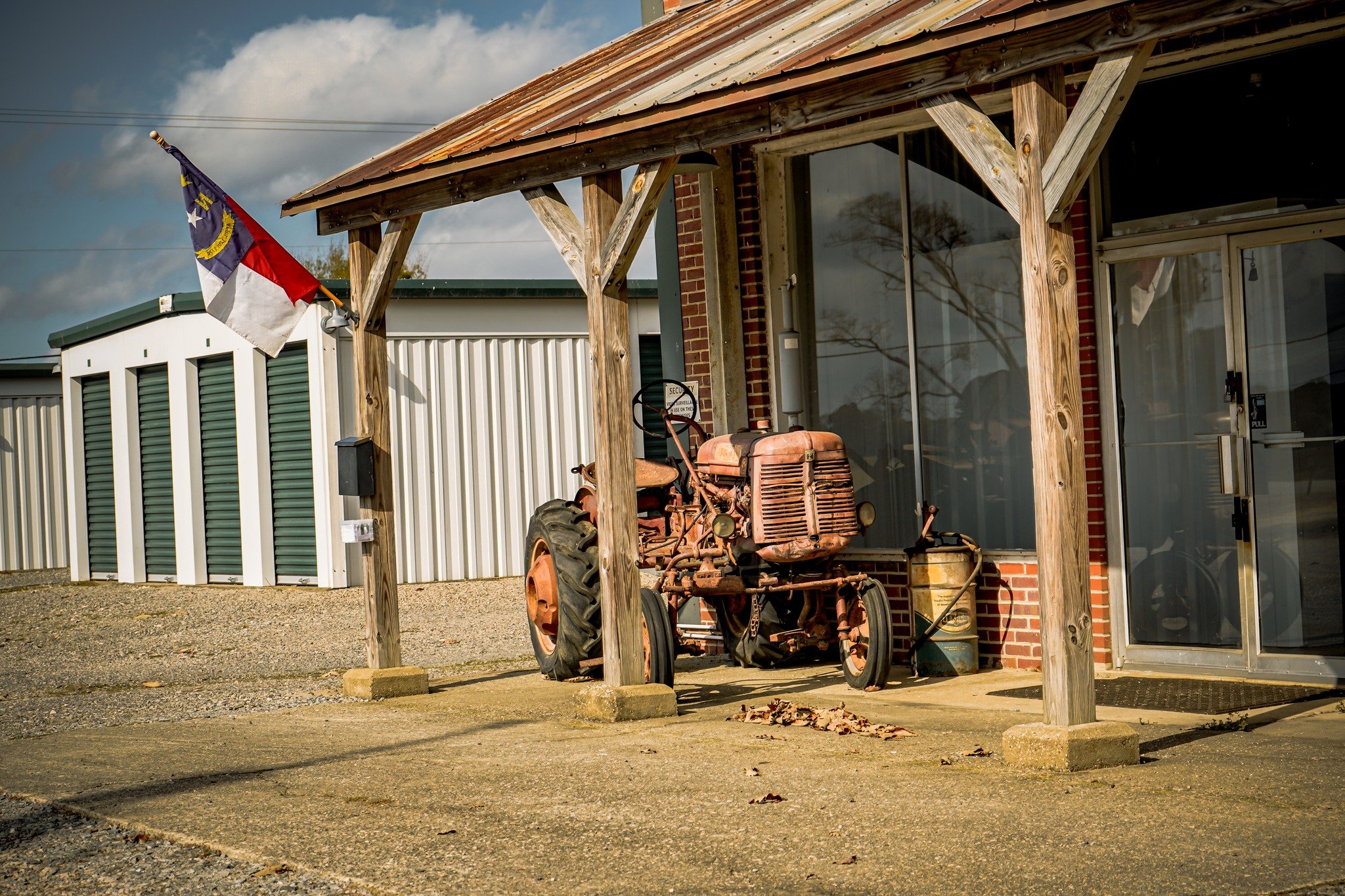 Old rusted tractor parked under wooden porch near brick building with glass door and window, North Carolina flag flying, gravel ground, cloudy sky.