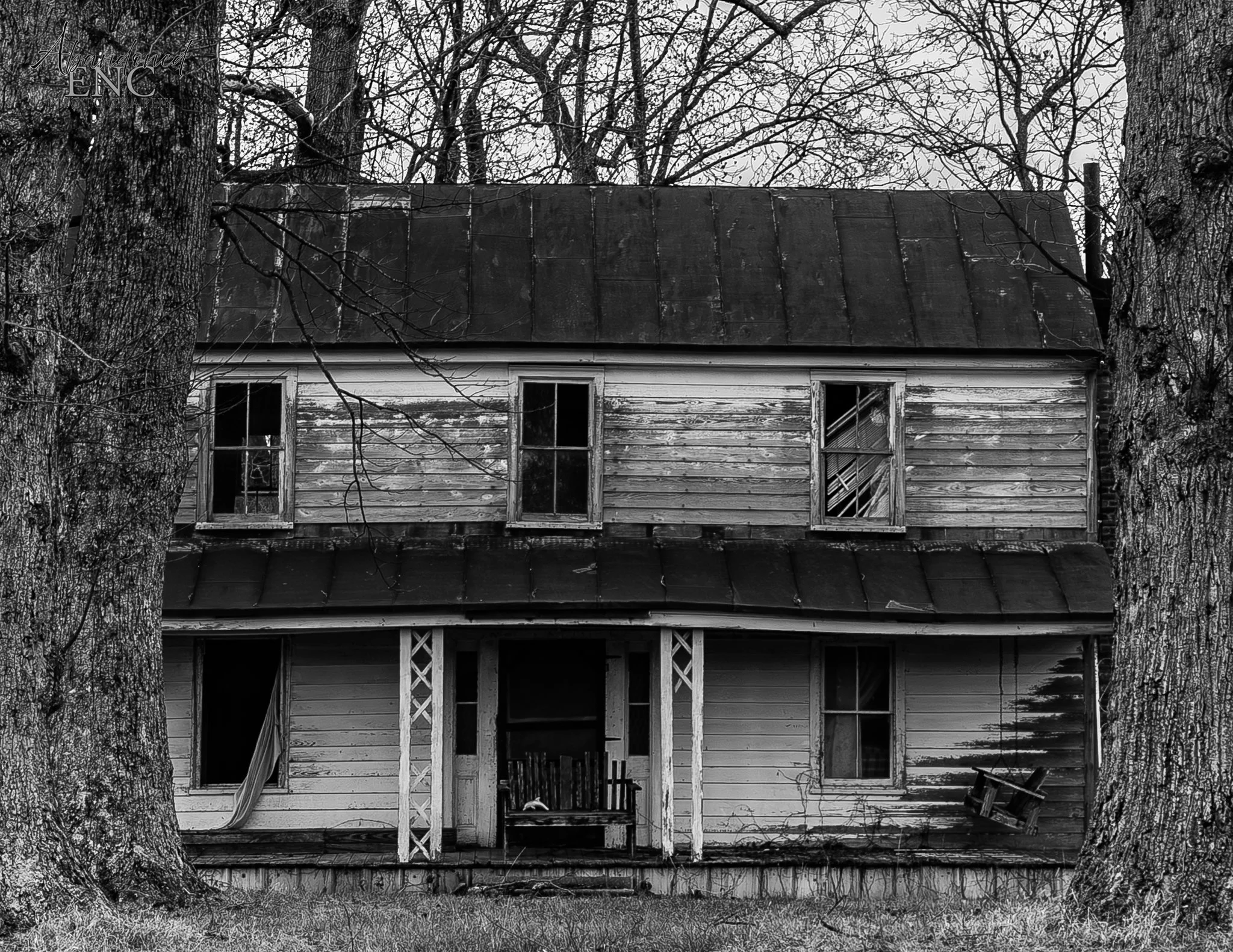 An old, abandoned house with peeling paint, broken windows, and a sagging roof, surrounded by leafless trees.