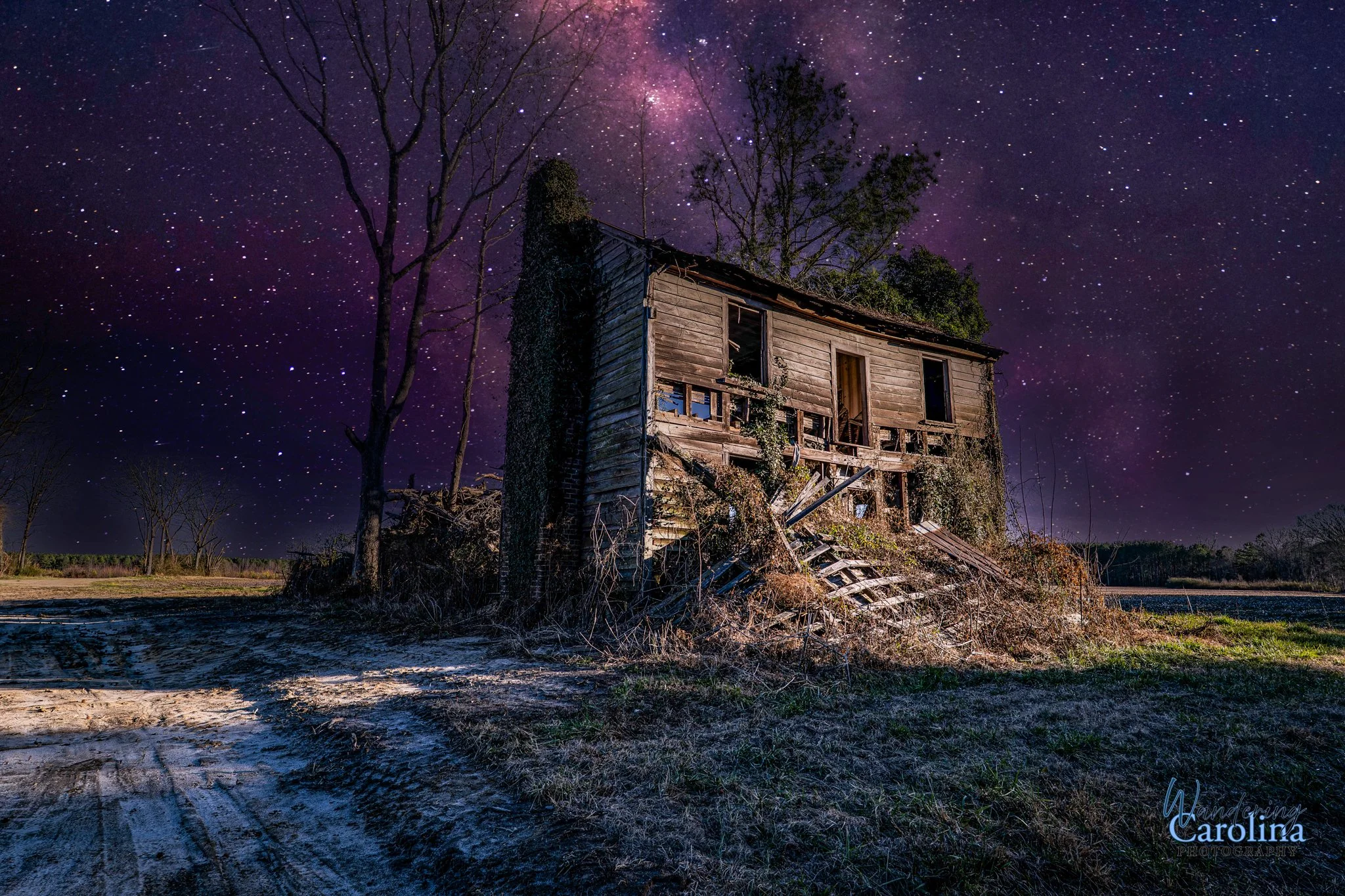 An abandoned, dilapidated wooden house on a dirt road at night under a starry sky with the Milky Way visible.