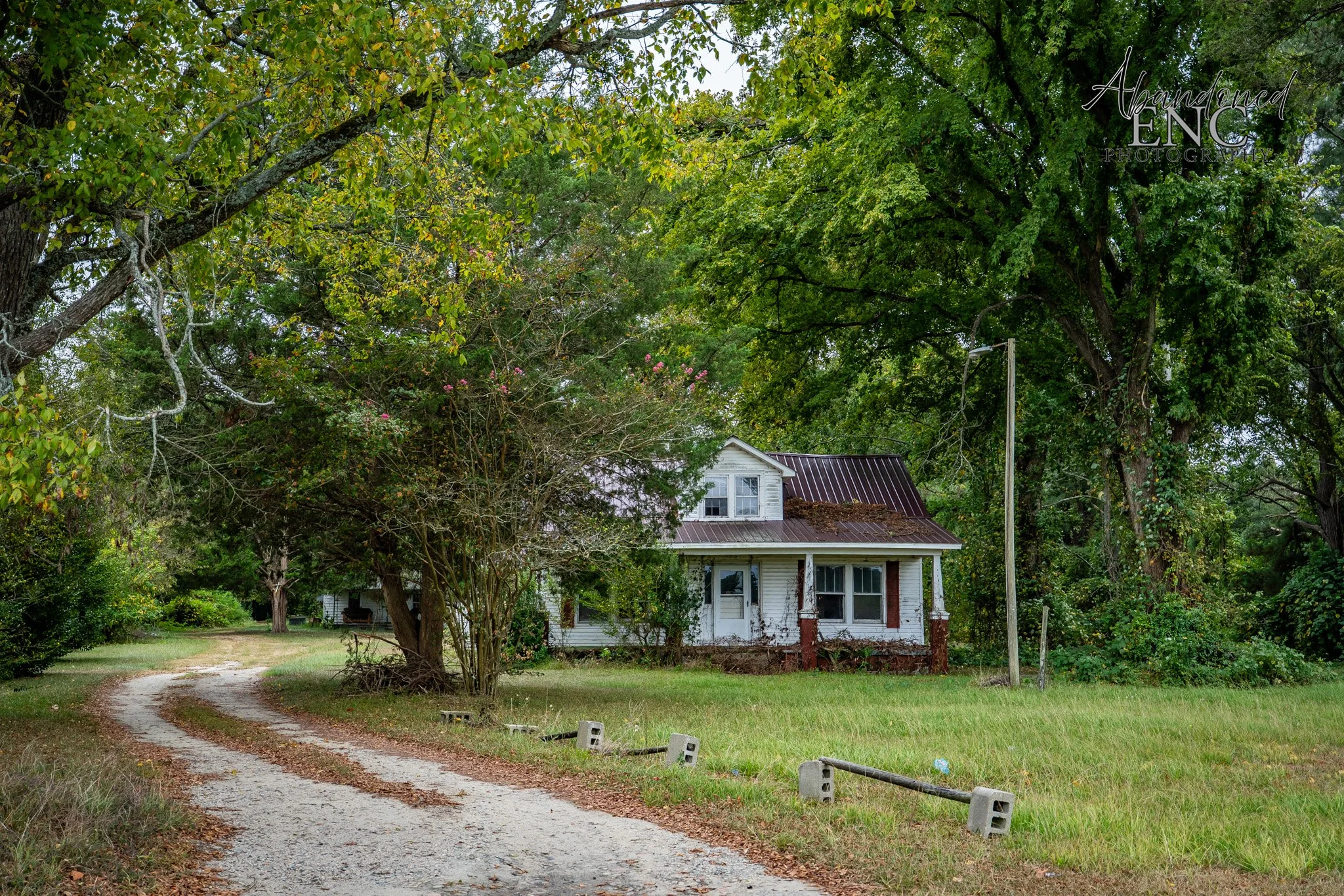 An old white house with a damaged roof, surrounded by tall green trees, and a gravel driveway with concrete barriers.