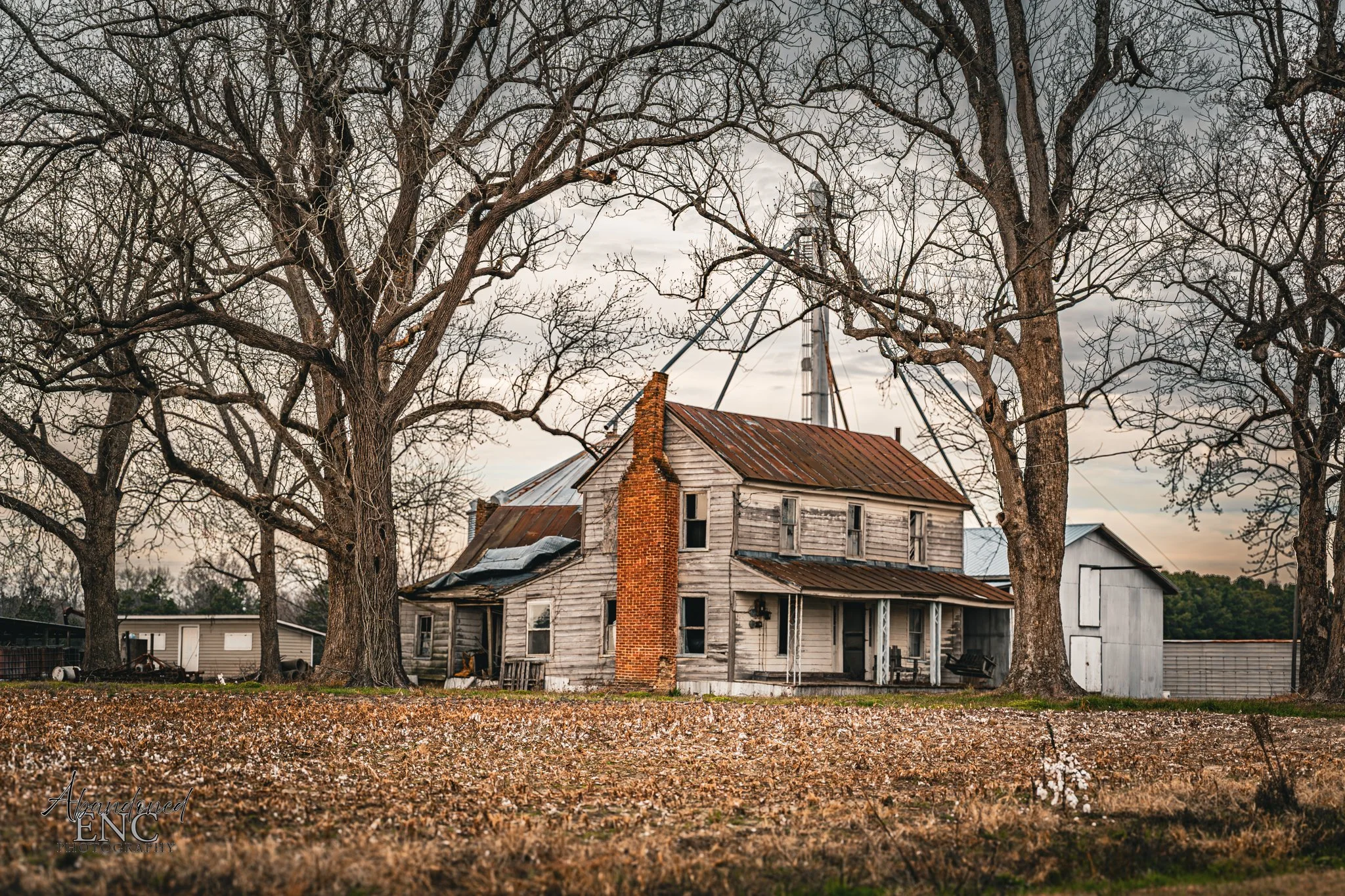 An old, weathered house with peeling paint and a rusted roof, surrounded by leafless trees, with a large antenna in the background and fallen leaves on the ground.