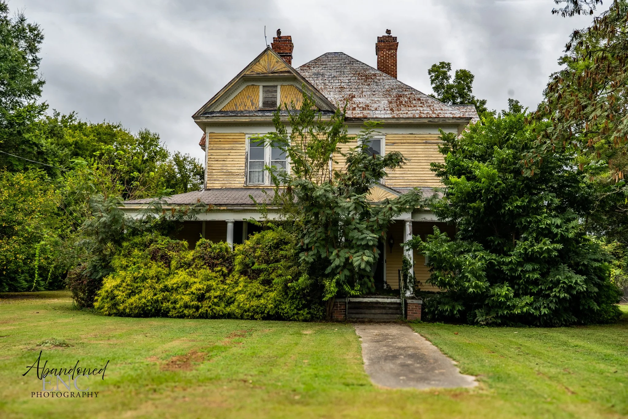 An old, abandoned two-story house with faded yellow paint, overgrown bushes, and large trees surrounding the front yard on a cloudy day.