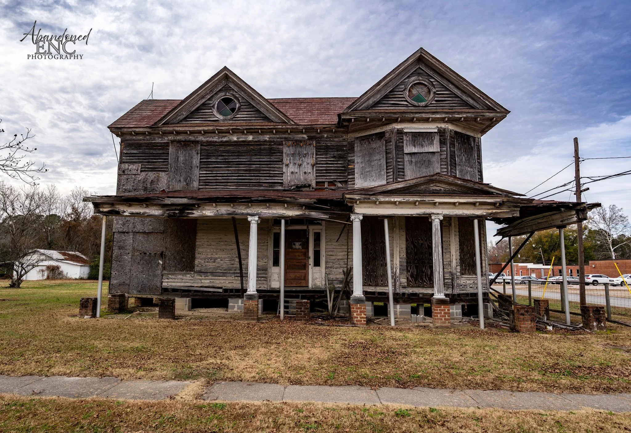 Old, dilapidated two-story wooden house with boarded-up windows and broken porch pillars, set on brick foundation blocks, under a cloudy sky.