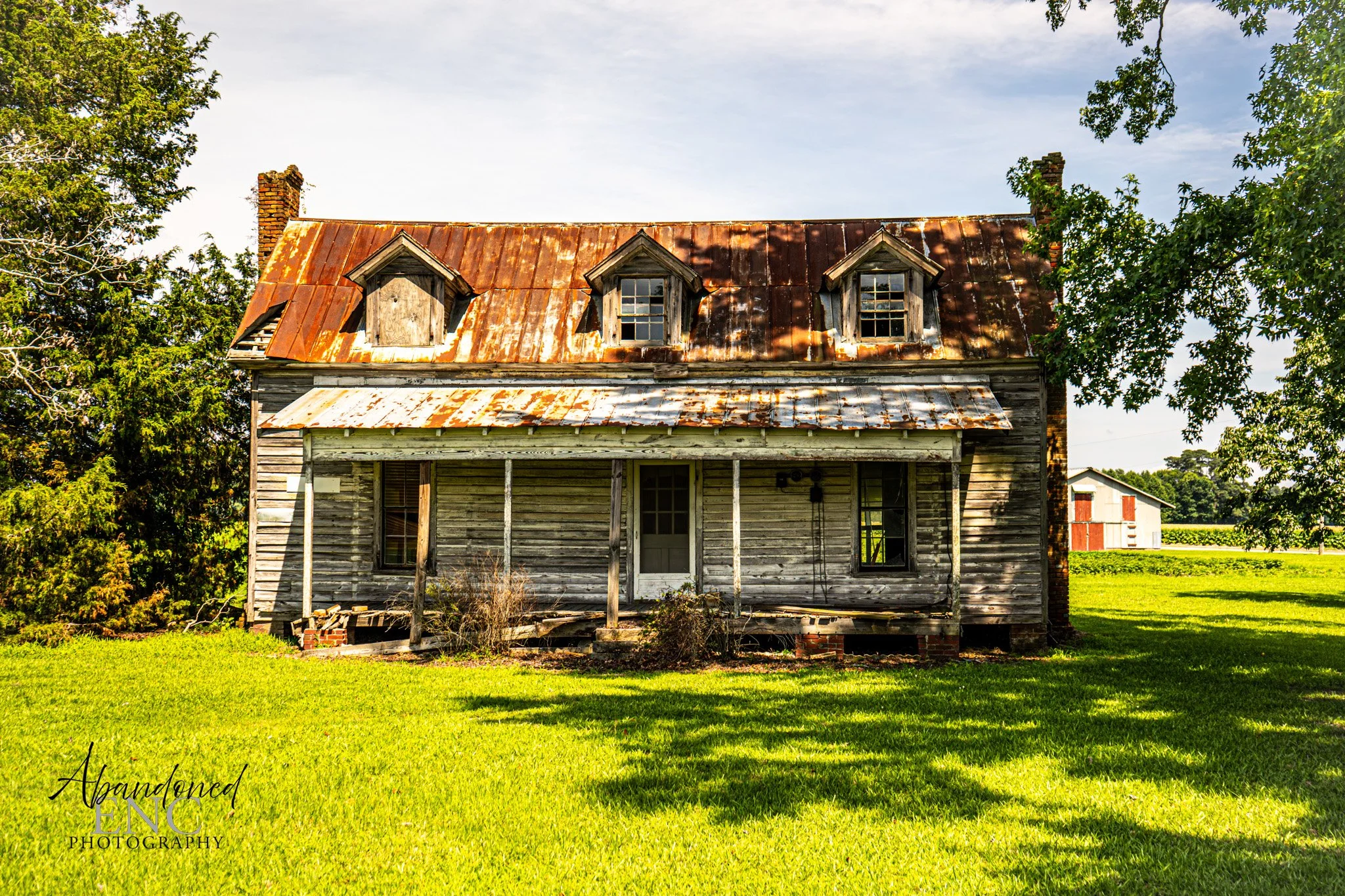 An old, abandoned house with a rusted metal roof, weathered wooden siding, and boarded-up windows, surrounded by green grass and trees.