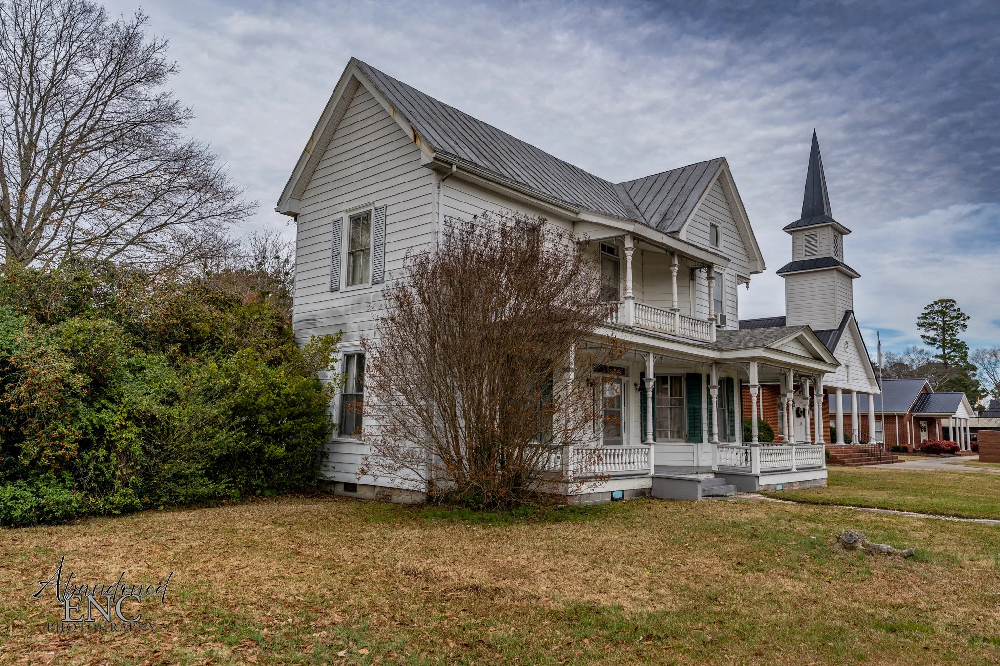 A large white Victorian house with a front porch and a steeple, surrounded by a grassy lawn and bushes, under a cloudy sky.