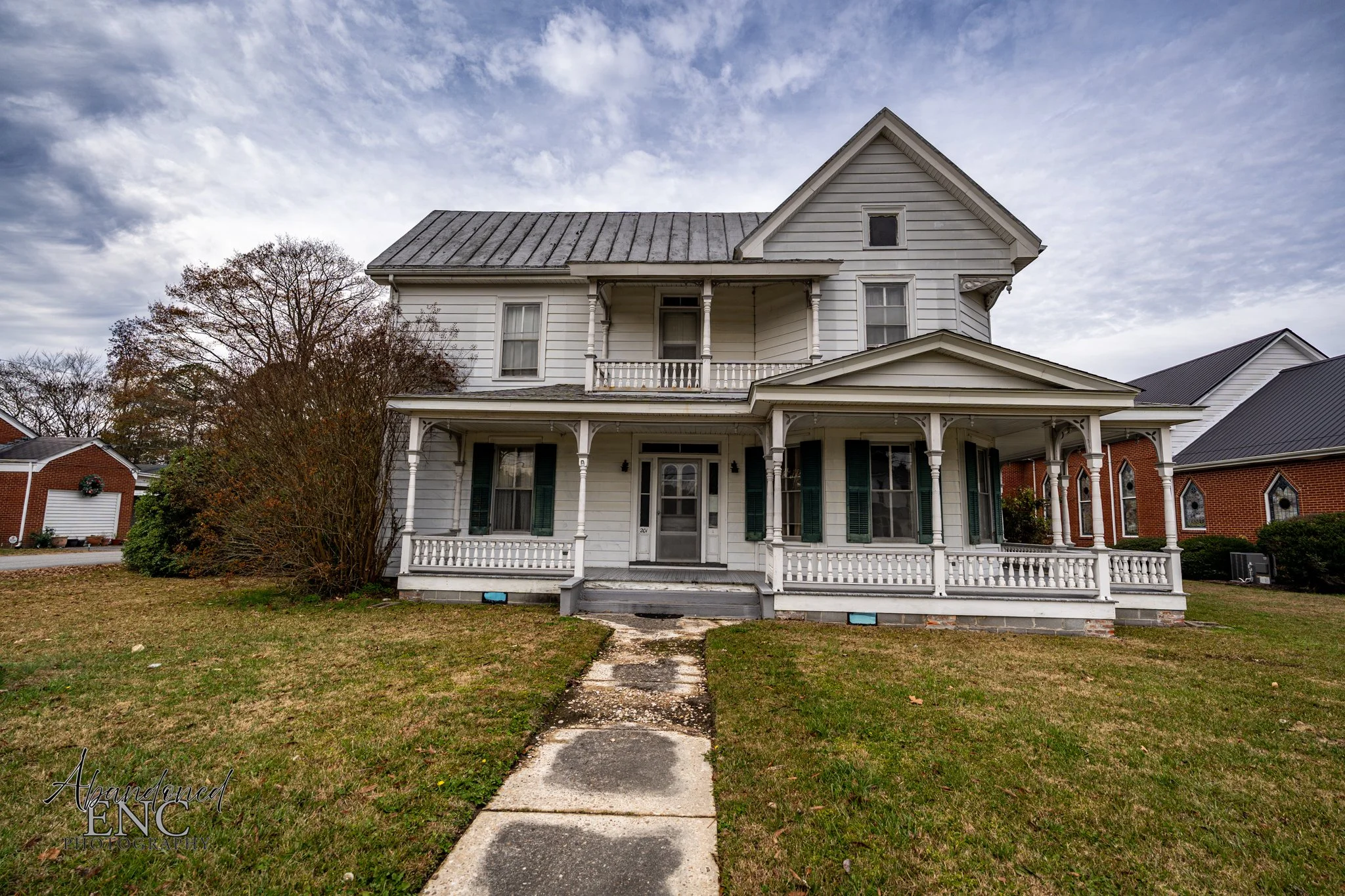 A two-story old white house with a front porch and green shutters in a residential neighborhood under a cloudy sky.