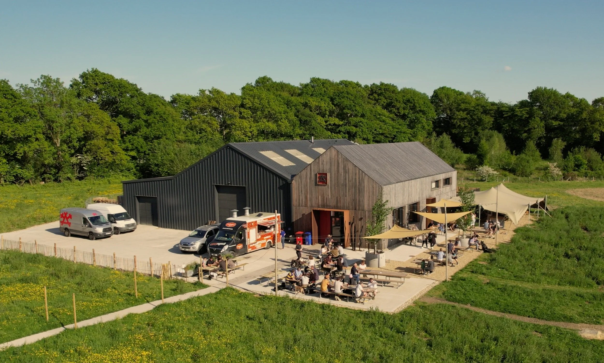 An outdoor food truck court adjacent to a barn with seating, umbrellas, and a parking lot with vehicles, surrounded by greenery. Taproom, Craft Beer, Street Food.