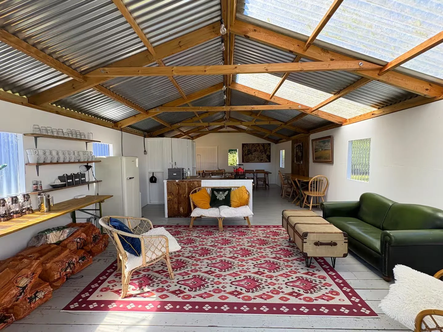 The image shows an open, rustic living room with a high, wooden beam and metal roof. The space is furnished with a green leather sofa, wicker chairs, a red patterned rug. Private Campsite Glamping Farm.